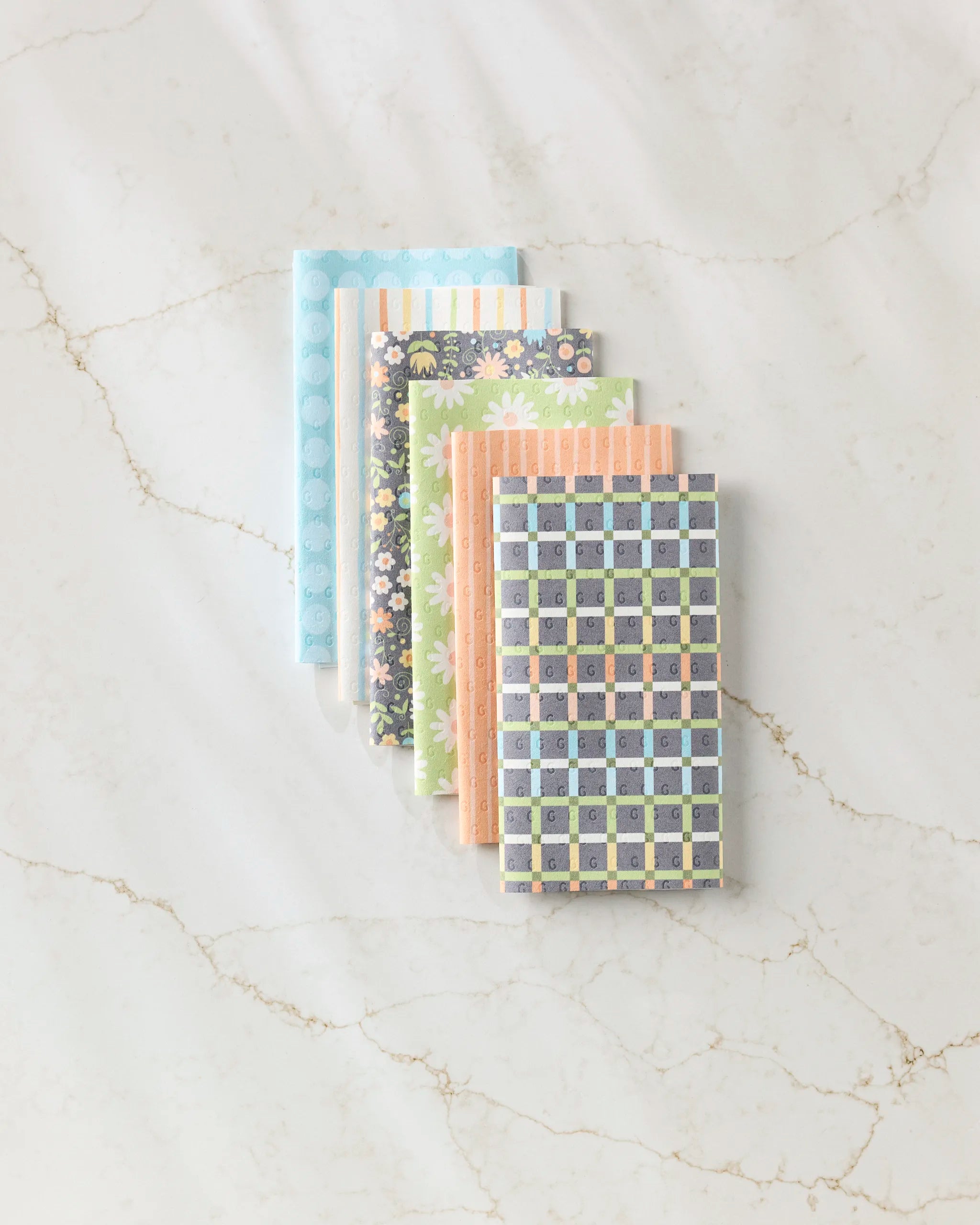 Six Mindfulnice "Mindful Medley" small notebooks, each with colorful floral, geometric, and stripe covers, are neatly arranged on a marble surface, evoking the vibrant look of eco-friendly kitchen towels.