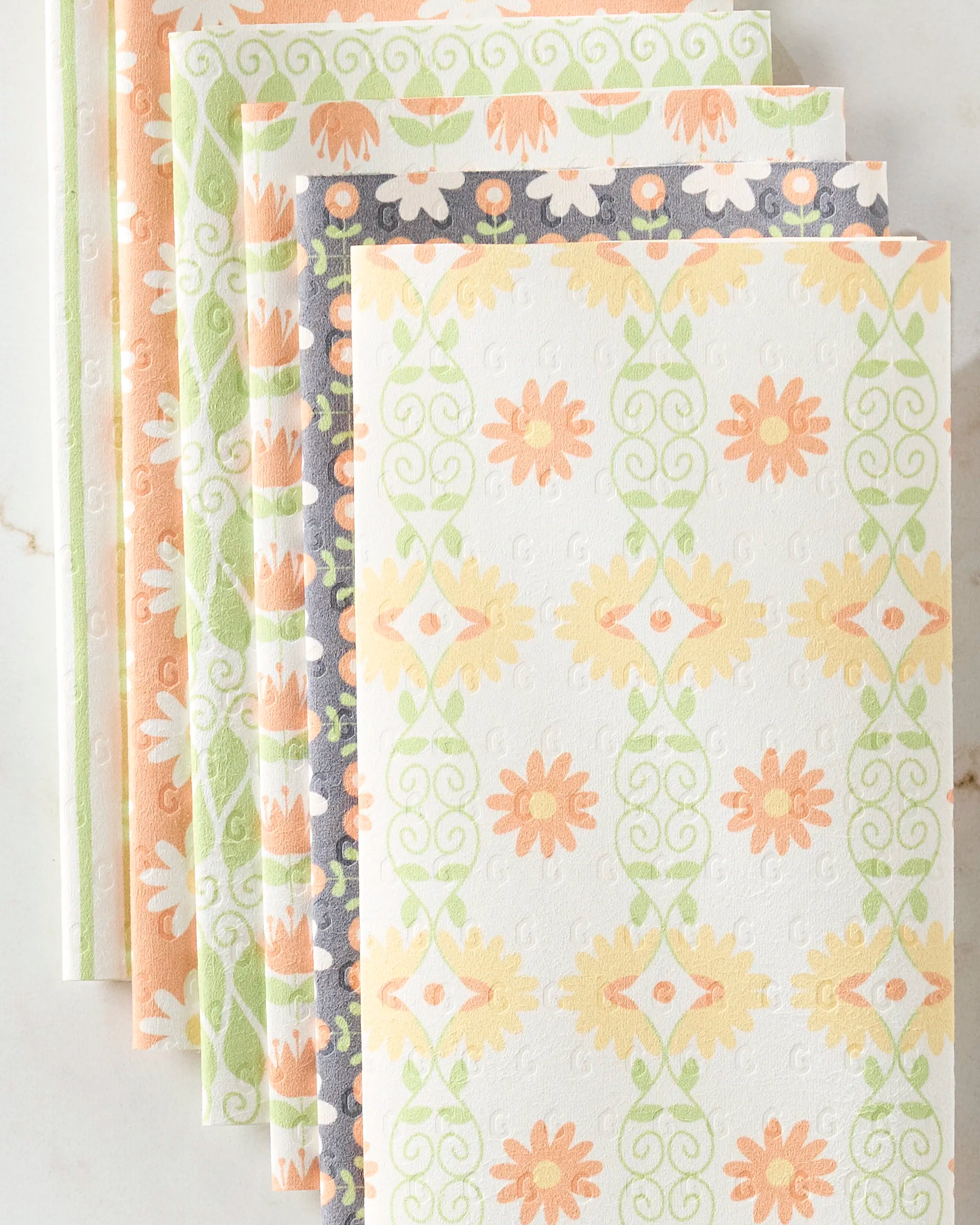 Five Mindfulnice Mindful Mix reusable paper towels are fanned out on a light marble surface, each featuring floral and geometric pastel designs in orange, green, peach, and cream.