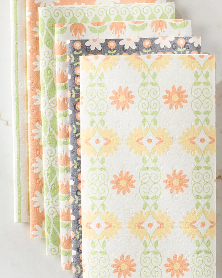 Five Mindfulnice Mindful Mix reusable paper towels are fanned out on a light marble surface, each featuring floral and geometric pastel designs in orange, green, peach, and cream.