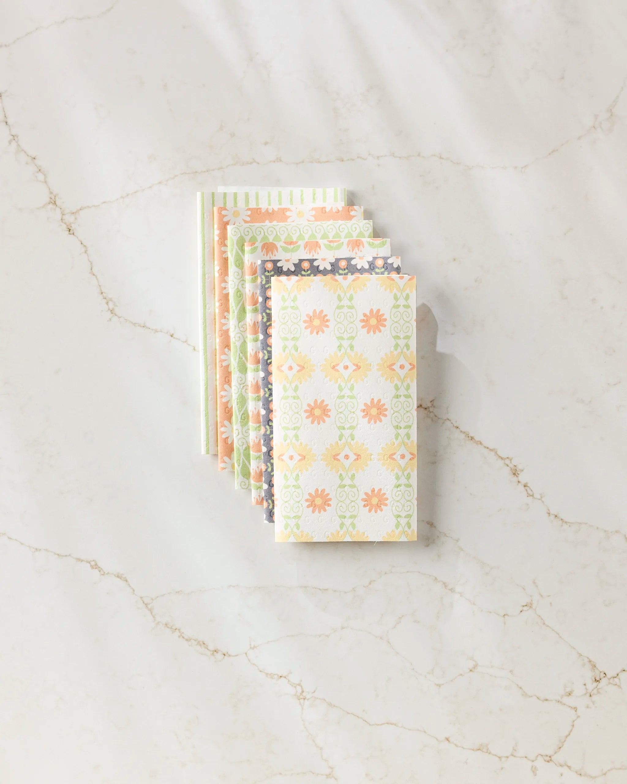 A stack of Mindful Mix colorful, patterned notebooks by Mindfulnice is fanned out on a white marble surface with veining, next to a neatly folded set of eco-friendly paper towels.