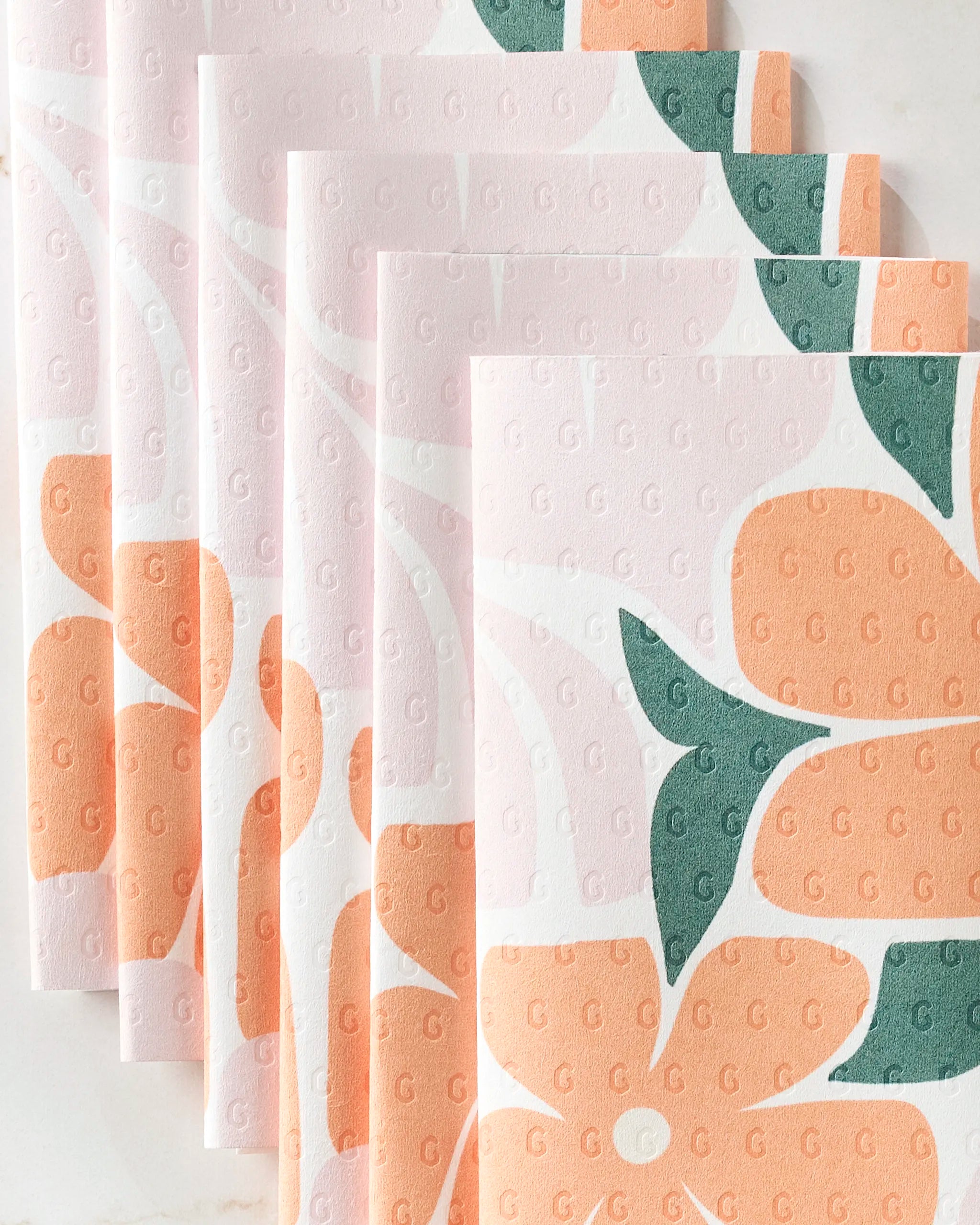 Five eco-friendly Lisa Nelson “Modern Floral” paper napkins overlap, featuring orange flowers and green leaves on pale pink, each embossed with a small “G” in a grid pattern.