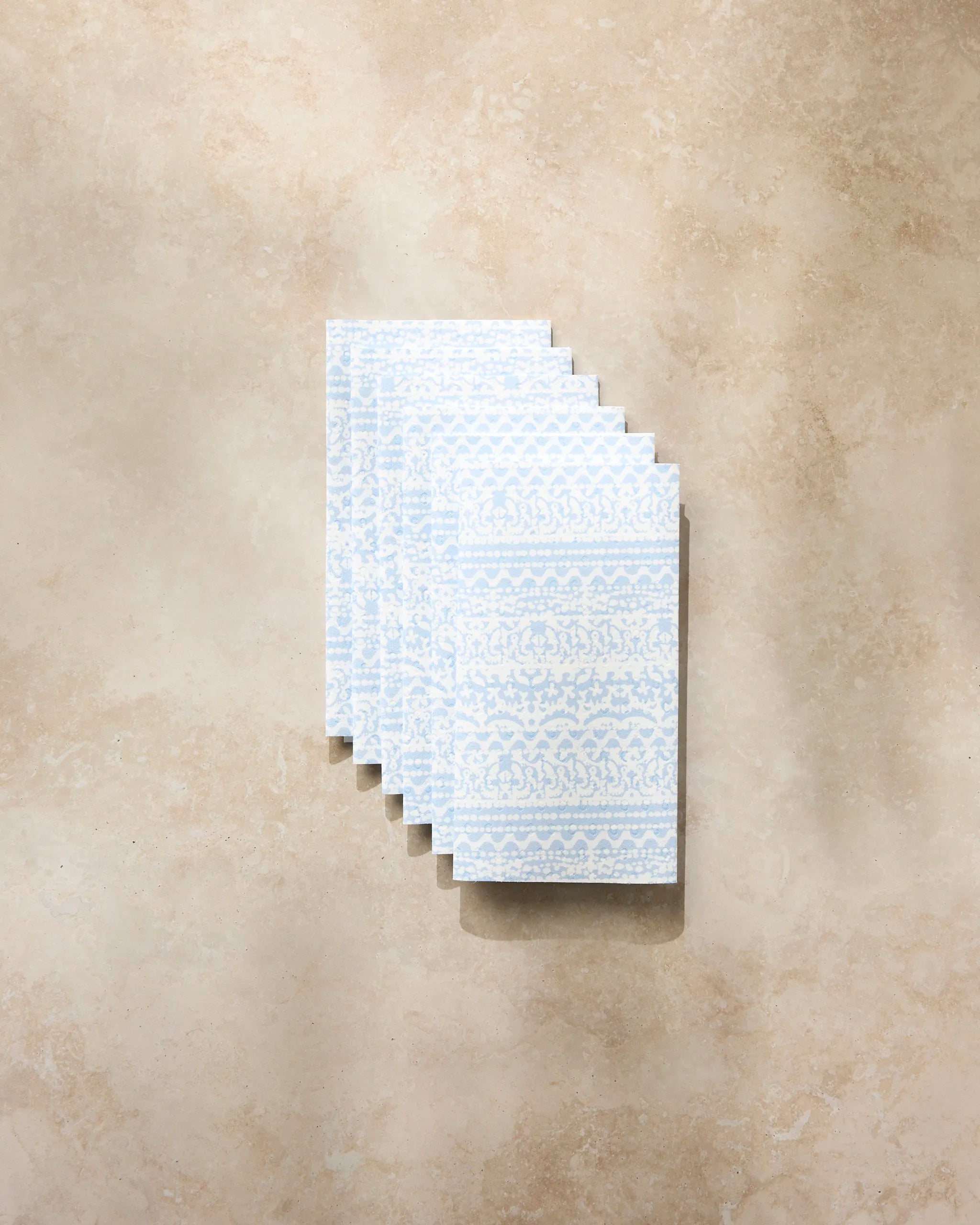 A stack of Geometry’s Modern Lace Not Paper Towels in light blue and white, neatly arranged on a beige stone surface, brings an eco-friendly accent to your kitchen.
