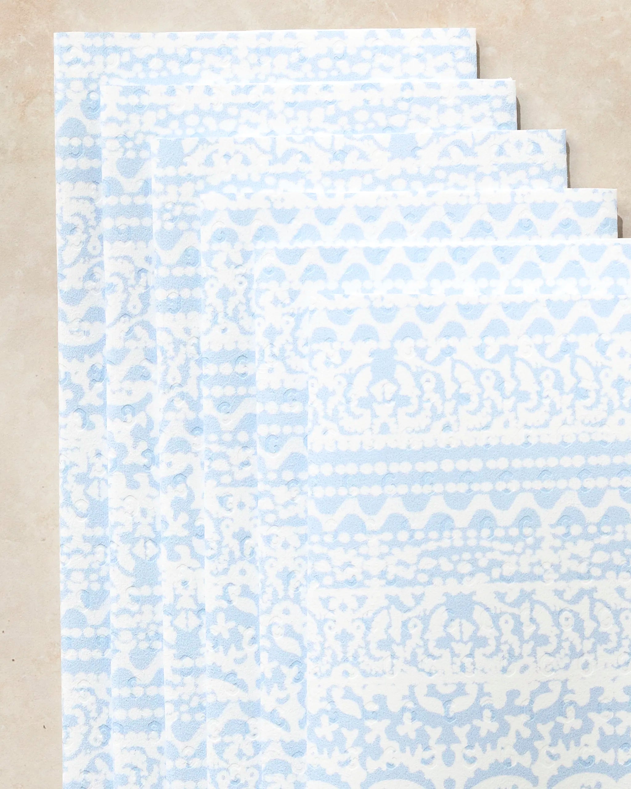 Five rectangular Modern Lace reusable paper towels by Geometry, featuring pale blue and white intricate patterns, are stacked in a slightly fanned arrangement on a light beige surface.
