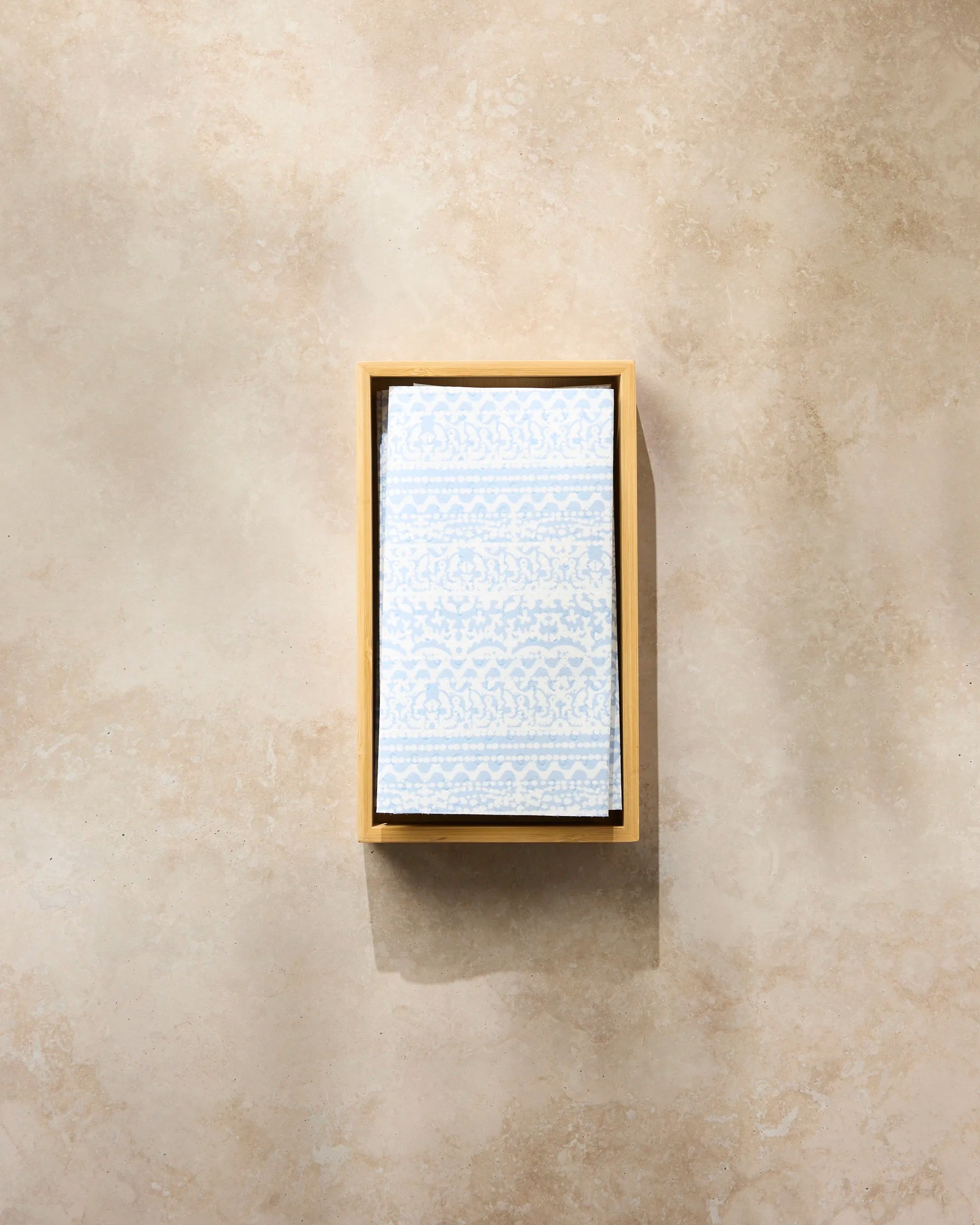 The Geometry Modern Lace reusable paper towel, featuring a light blue and white pattern, is neatly folded in a rectangular wooden tray on a beige marble surface.