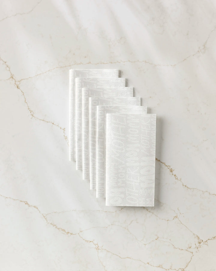 Seven white textured "Mom" reusable paper towels by GEOMETRY, featuring embossed text, are neatly stacked in an overlapping arrangement on a white marble surface with light brown veins.