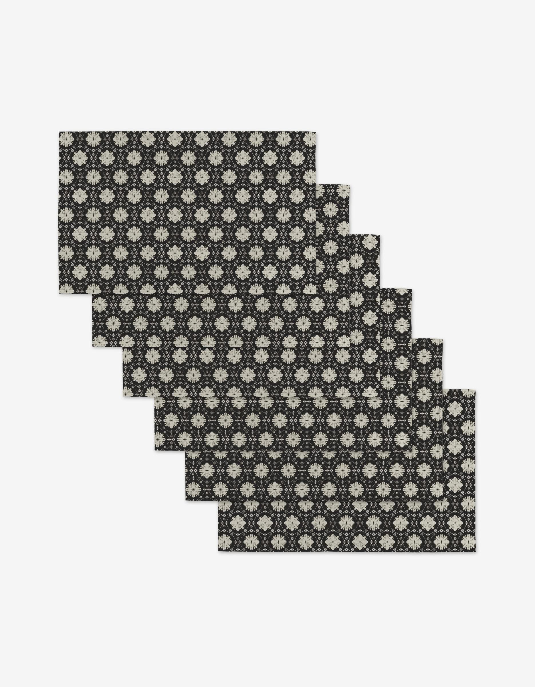 Six Monochrome Florals placemats by Danelys Sidron, rectangular and black with a white floral pattern, made from post-consumer recycled materials, are shown in a staggered stack on a white background.