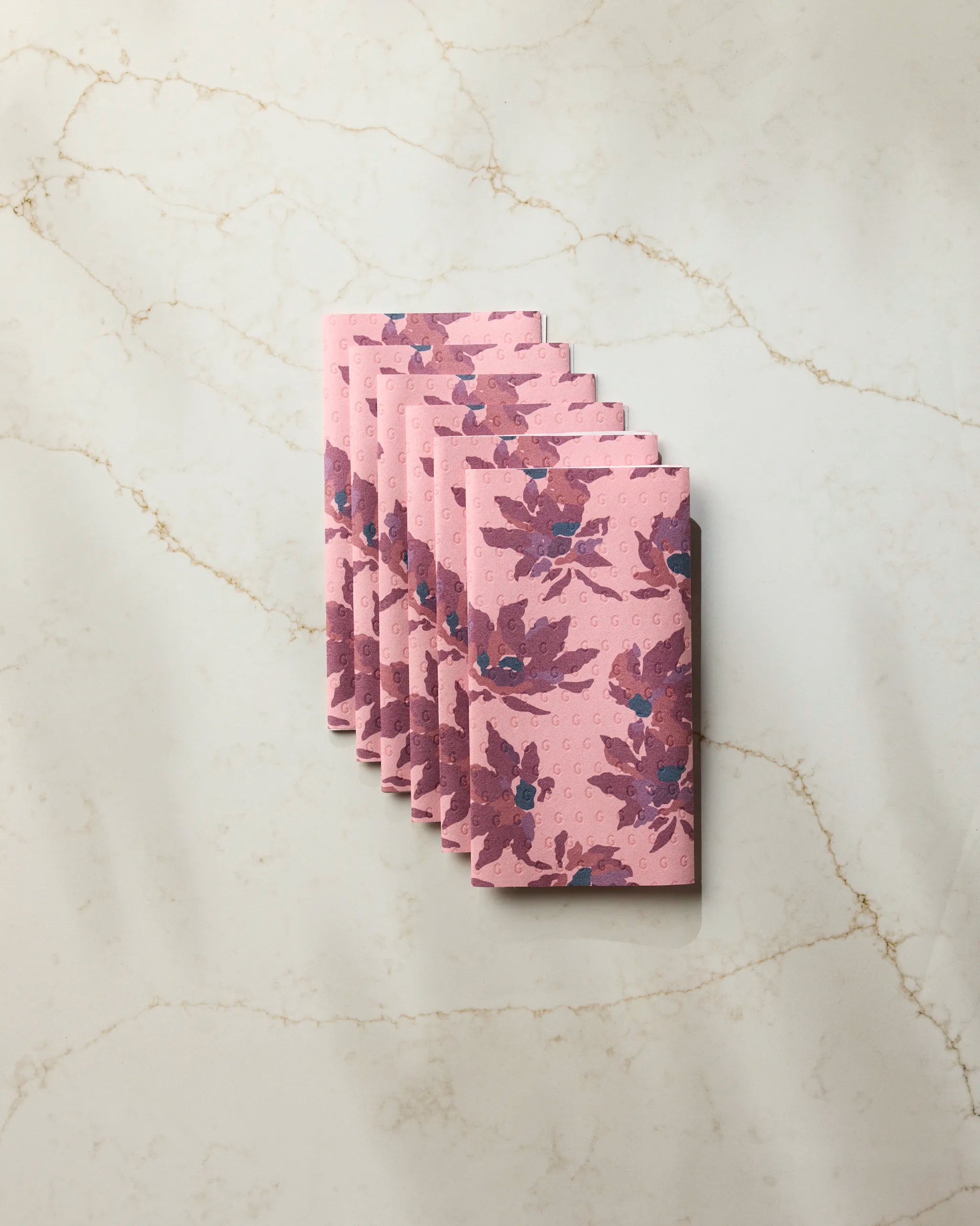 Seven neatly folded Mulberry Bloom reusable towels by Geometry, featuring a pink base and purple floral pattern, are stacked on light marble with gold veining—an elegant, eco-friendly alternative to paper towels.