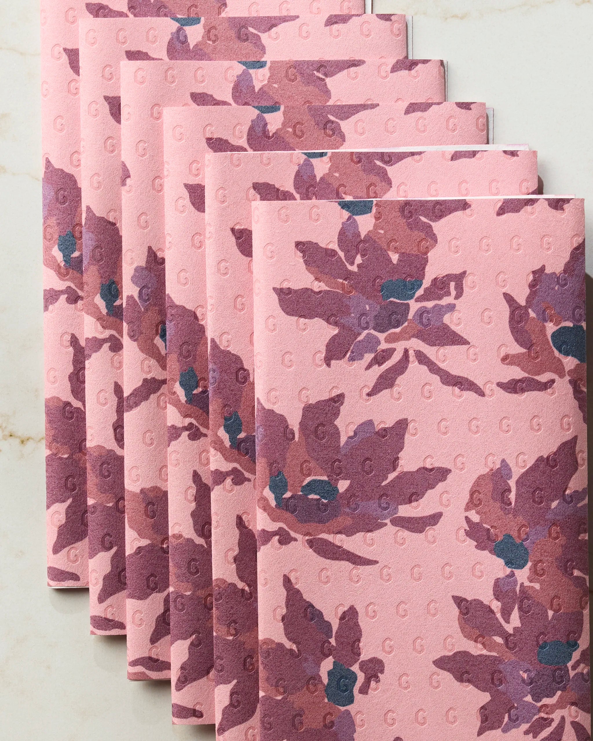 Six Mulberry Bloom napkins from Geometry feature a purple floral design with subtle G letters, neatly stacked on marble—ideal as eco-friendly kitchen towels or reusable paper towels for any stylish table setting.