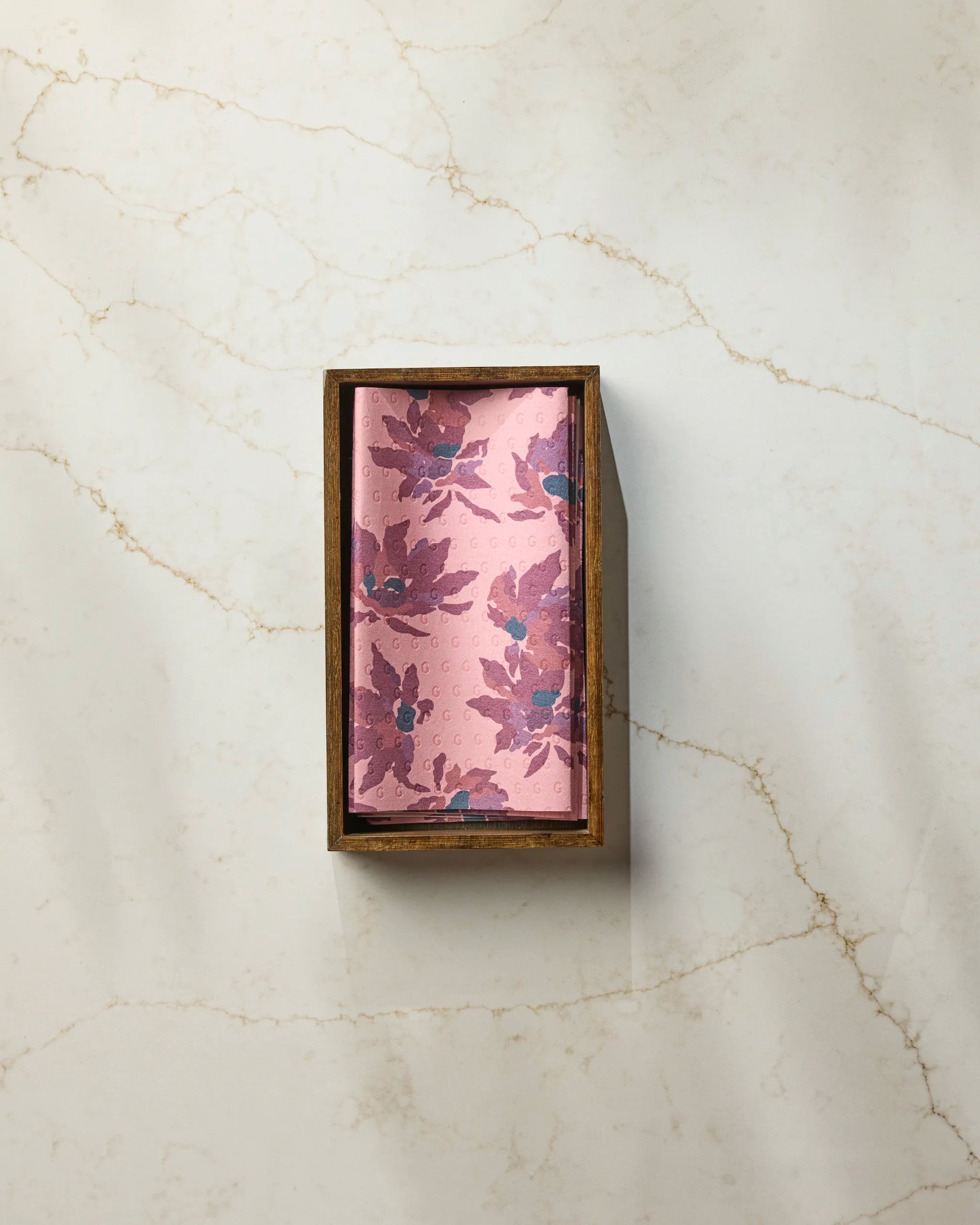 A Mulberry Bloom reusable paper towel by Geometry, featuring a pink floral pattern, is placed in a rectangular box on a white marble surface with light brown veins.