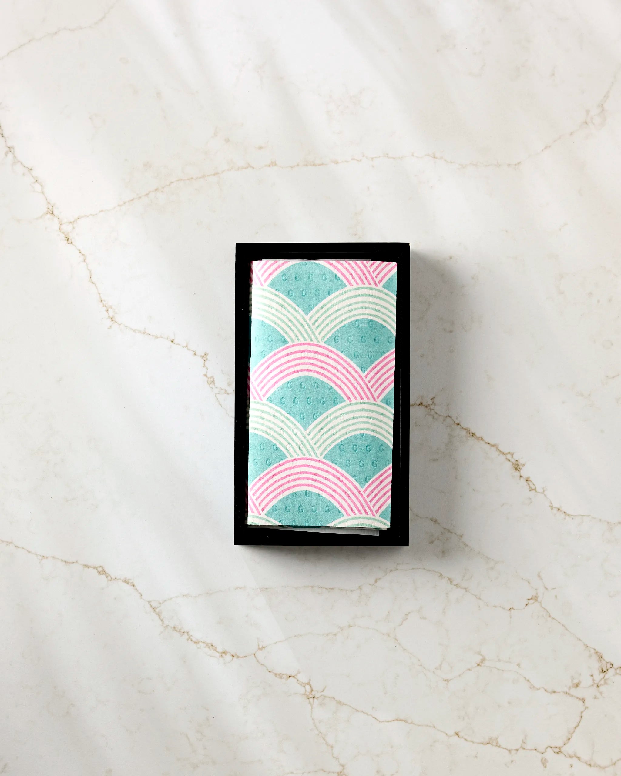 The Nalin bar by Amy MacCready, wrapped in pastel blue and pink patterned recycled paper towels, rests in a black tray on a light marble surface.
