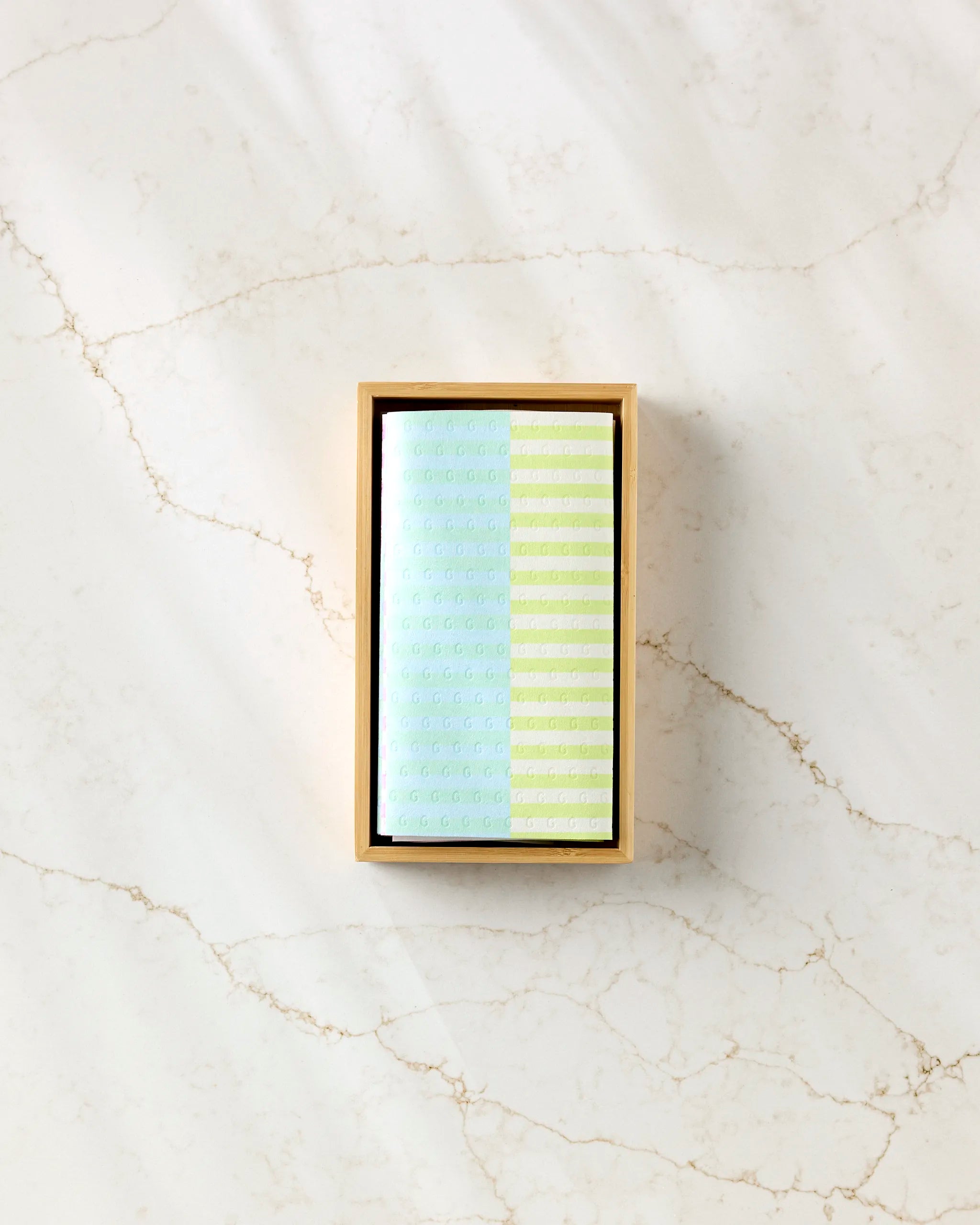 A rectangular box labeled "Neon Nights" by GEOMETRY, wrapped in light blue and green patterns, sits on a white marble surface with subtle gold veining, hinting at eco-friendly kitchen towels inside.