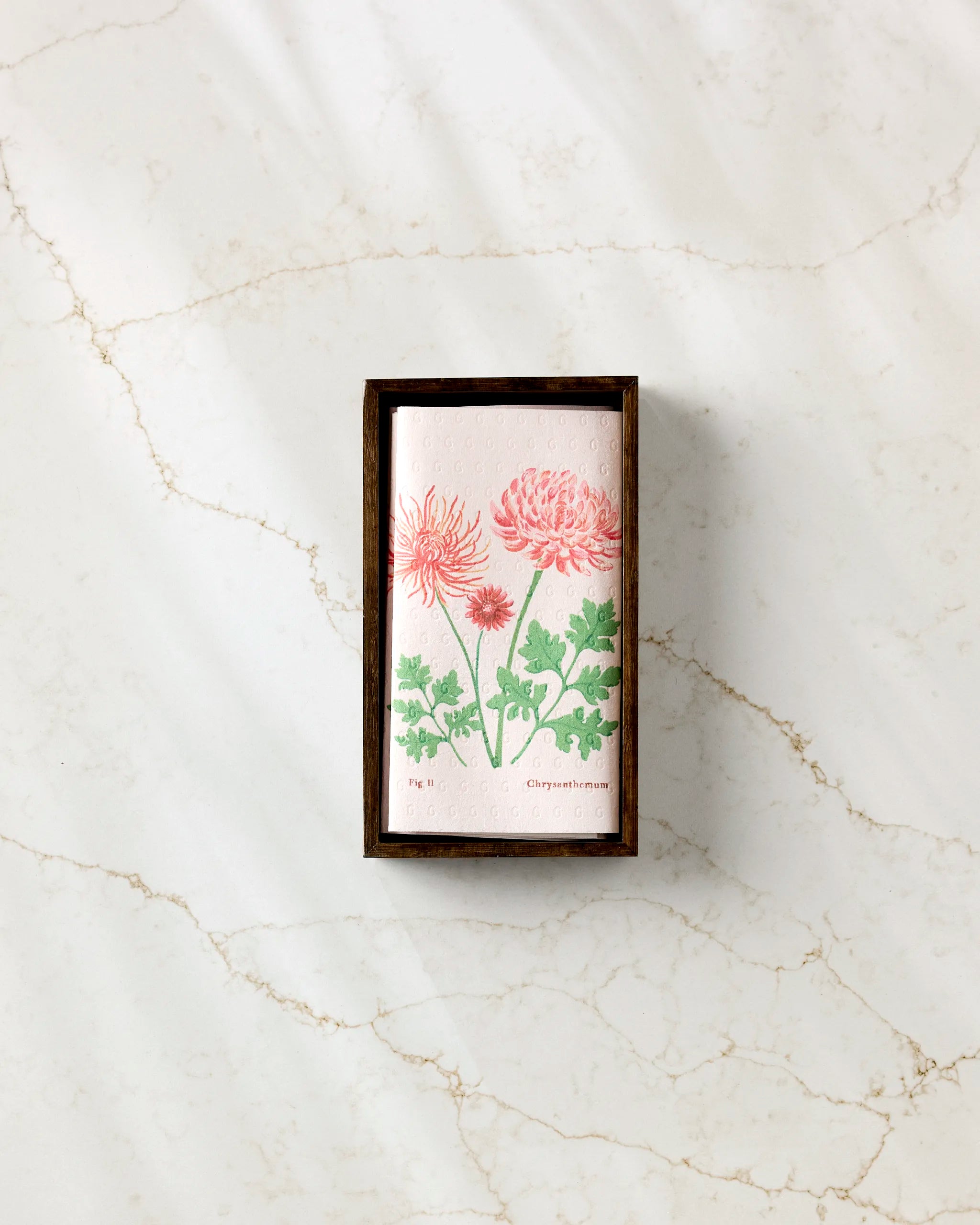 The Linda Fox "November Chrysanthemum" soap, featuring pink chrysanthemum illustrations, is displayed in a small rectangular wooden box on white marble with light brown veins, beside neatly folded eco-friendly kitchen towels.