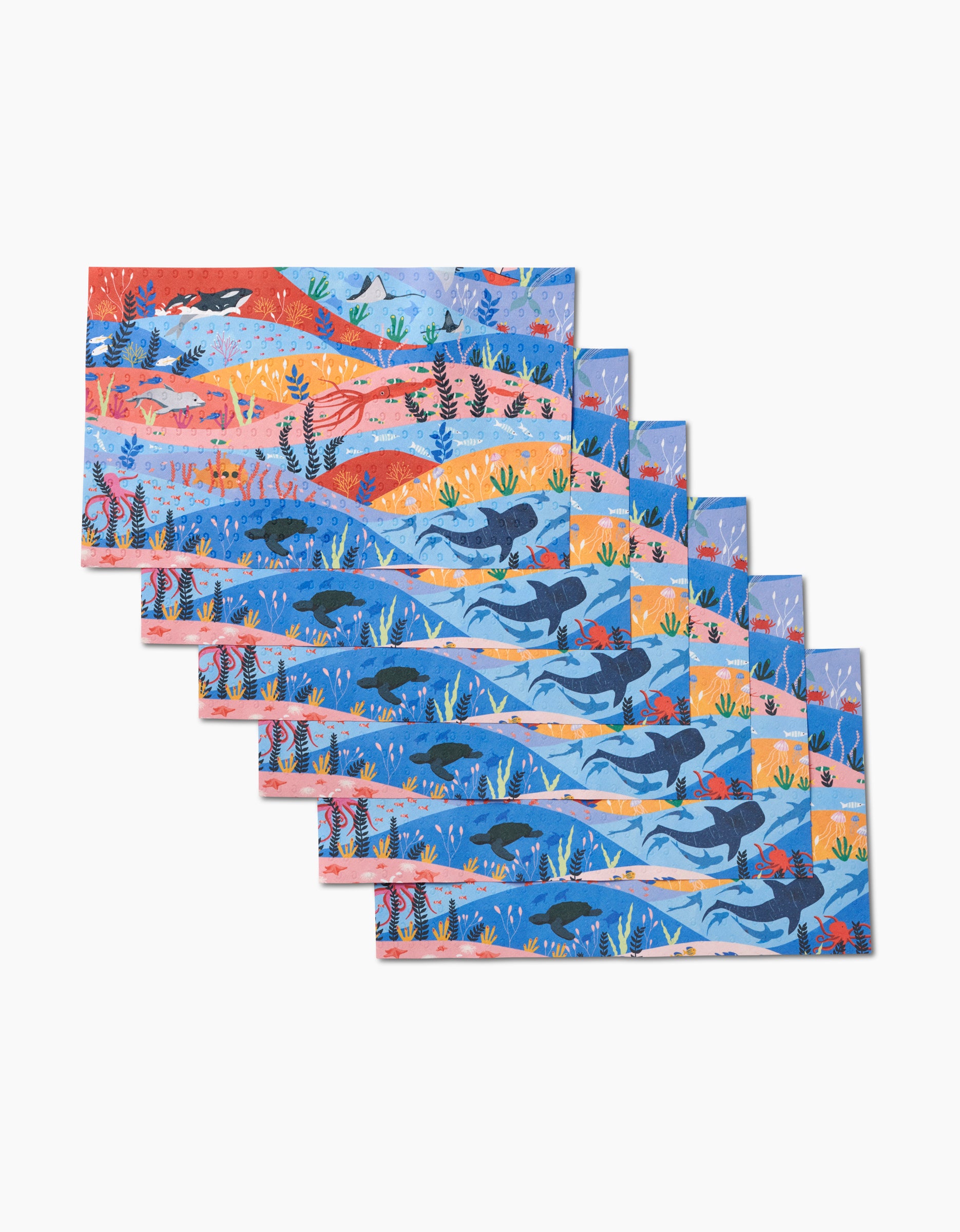 Oceans by Ceyda Alasar features seven illustrated placemats overlapping in a fan shape, each showcasing vibrant ocean scenes with whales, fish, and sea plants on white, made from eco-friendly post-consumer recycled materials.