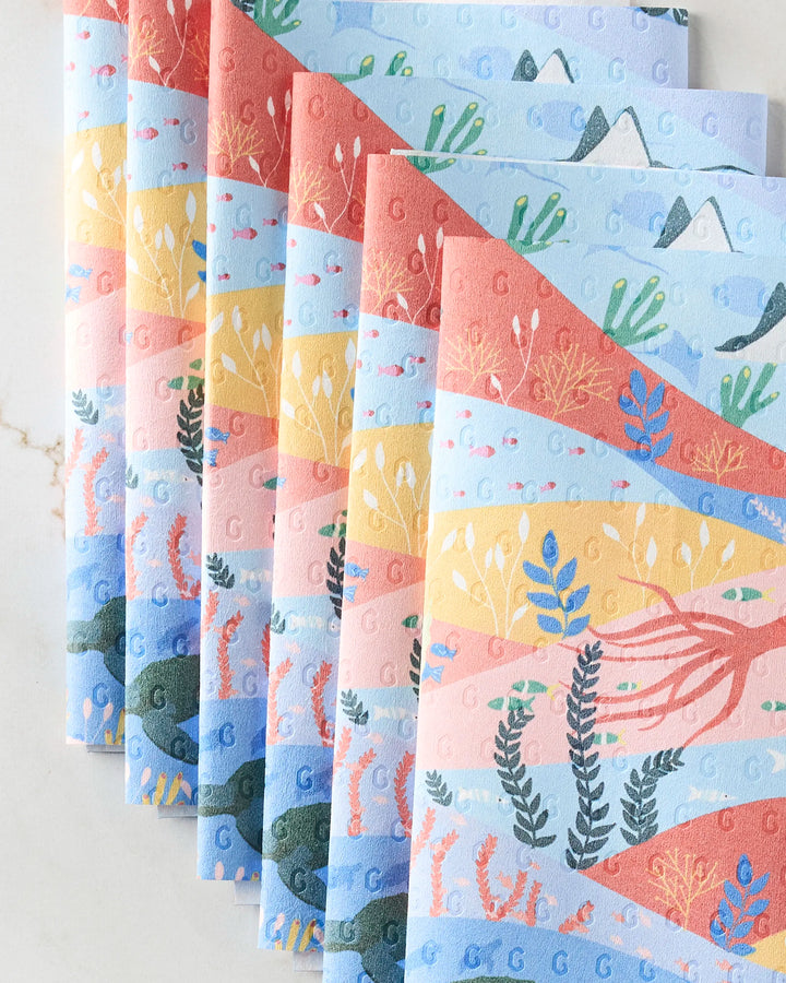 Oceans by Ceyda Alasar: eco-friendly paper towels in soft pastel blue, pink, and orange with illustrated underwater and desert scenes—fish, sea plants, coral, mountains, and cacti—artfully arranged in a fan pattern.