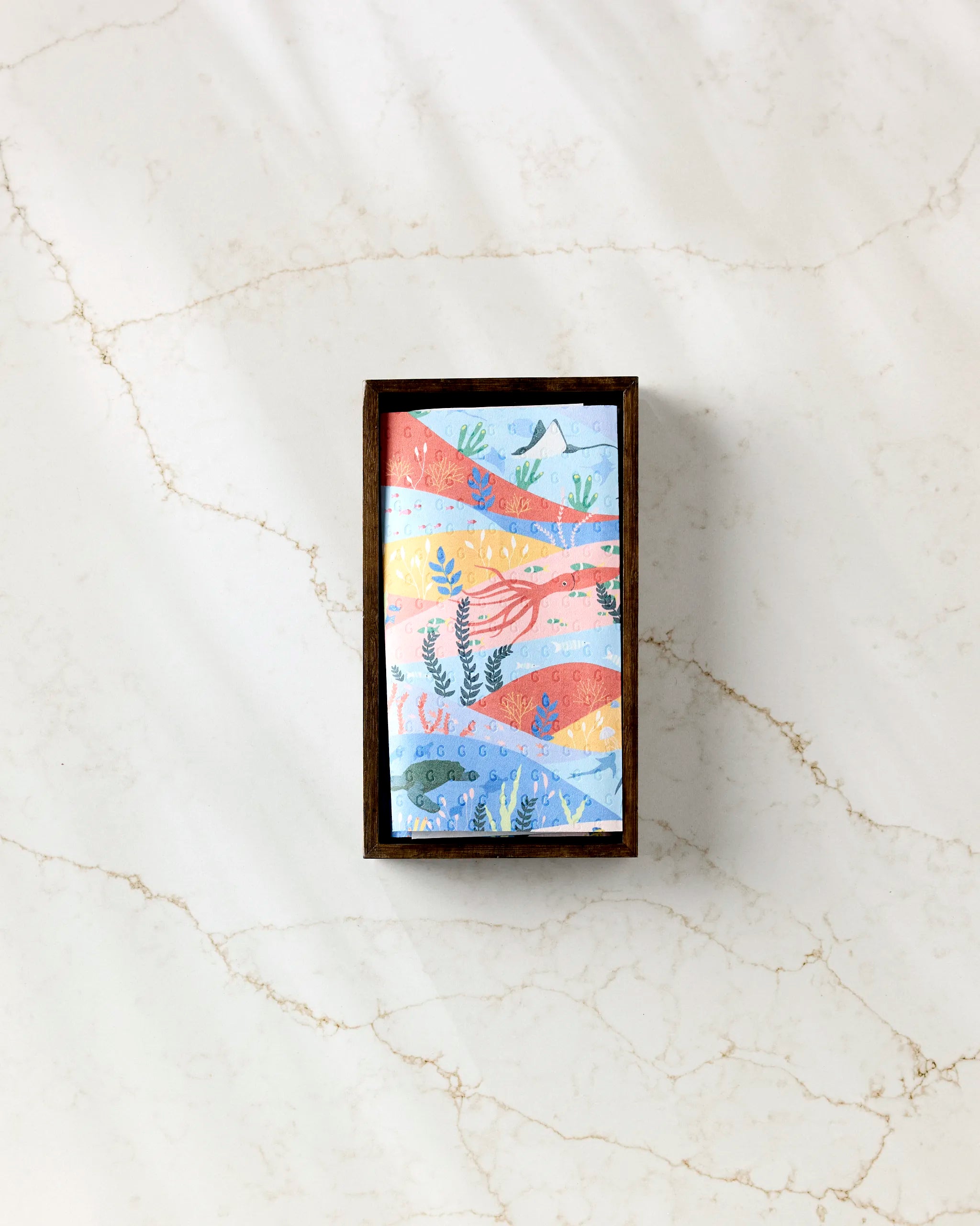 Oceans" by Ceyda Alasar—a rectangular wooden frame with colorful wavy botanical art in red, blue, yellow, and green—is displayed on white marble alongside eco-friendly paper towels.