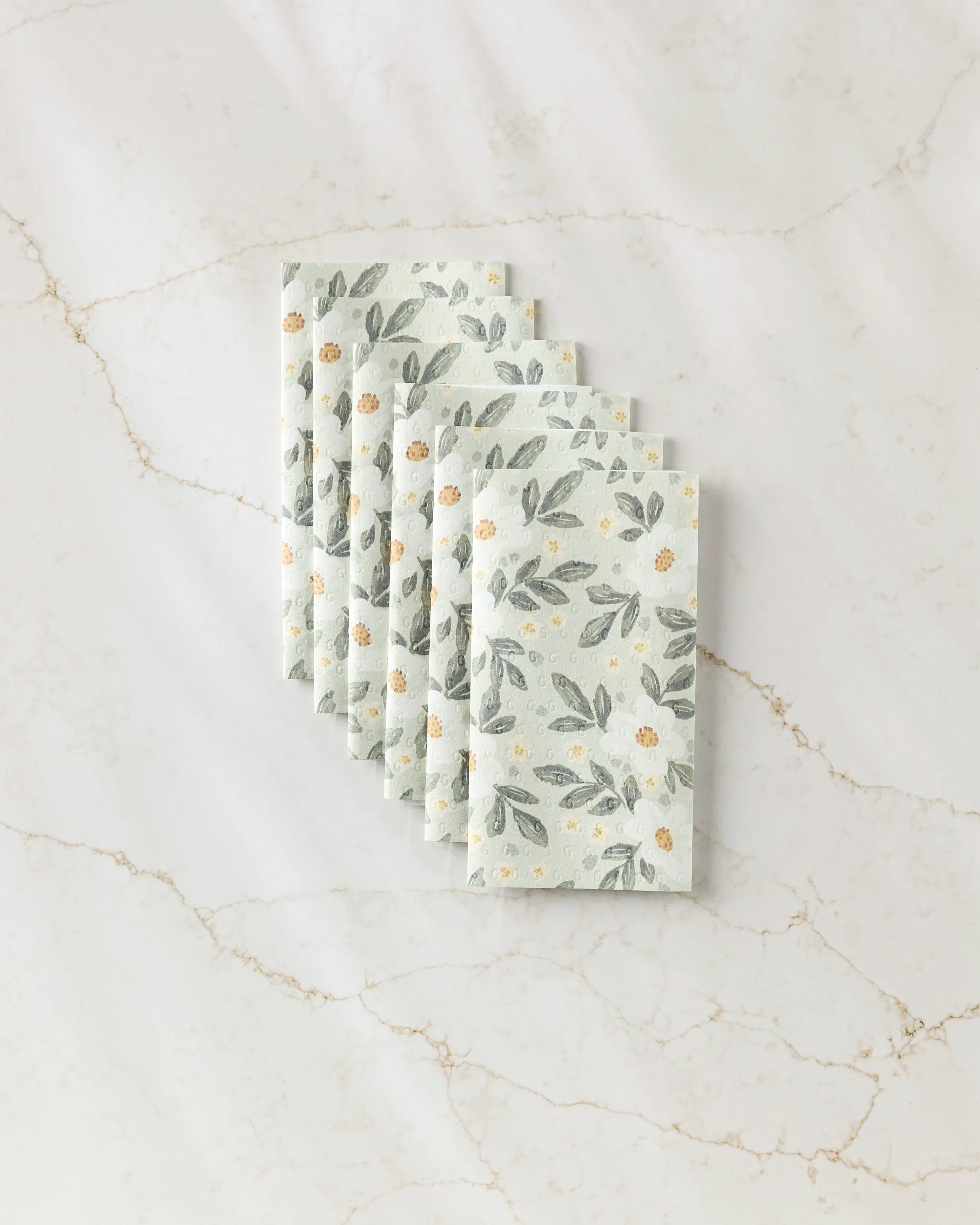 Six folded, reusable paper towels from Teri the Butter’s “Painted Spring Flowers” collection are displayed in a fan shape on a white marble surface with light brown veining.