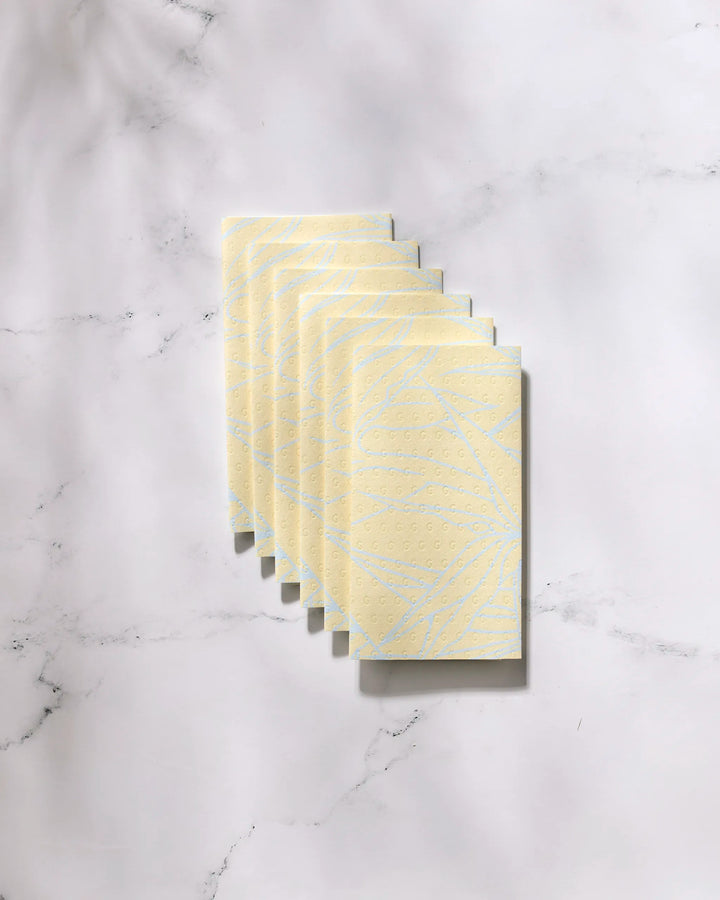 Six yellow eco-friendly GEOMETRY Paradise Blooms paper towels, featuring a subtle leaf and dotted pattern, are stacked diagonally on a white marble surface.