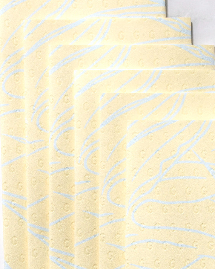 Five overlapping sheets of GEOMETRY's Paradise Blooms eco-friendly paper towels, featuring yellow textured surfaces with subtle blue wavy lines and small embossed G letters, are arranged diagonally on a light background.