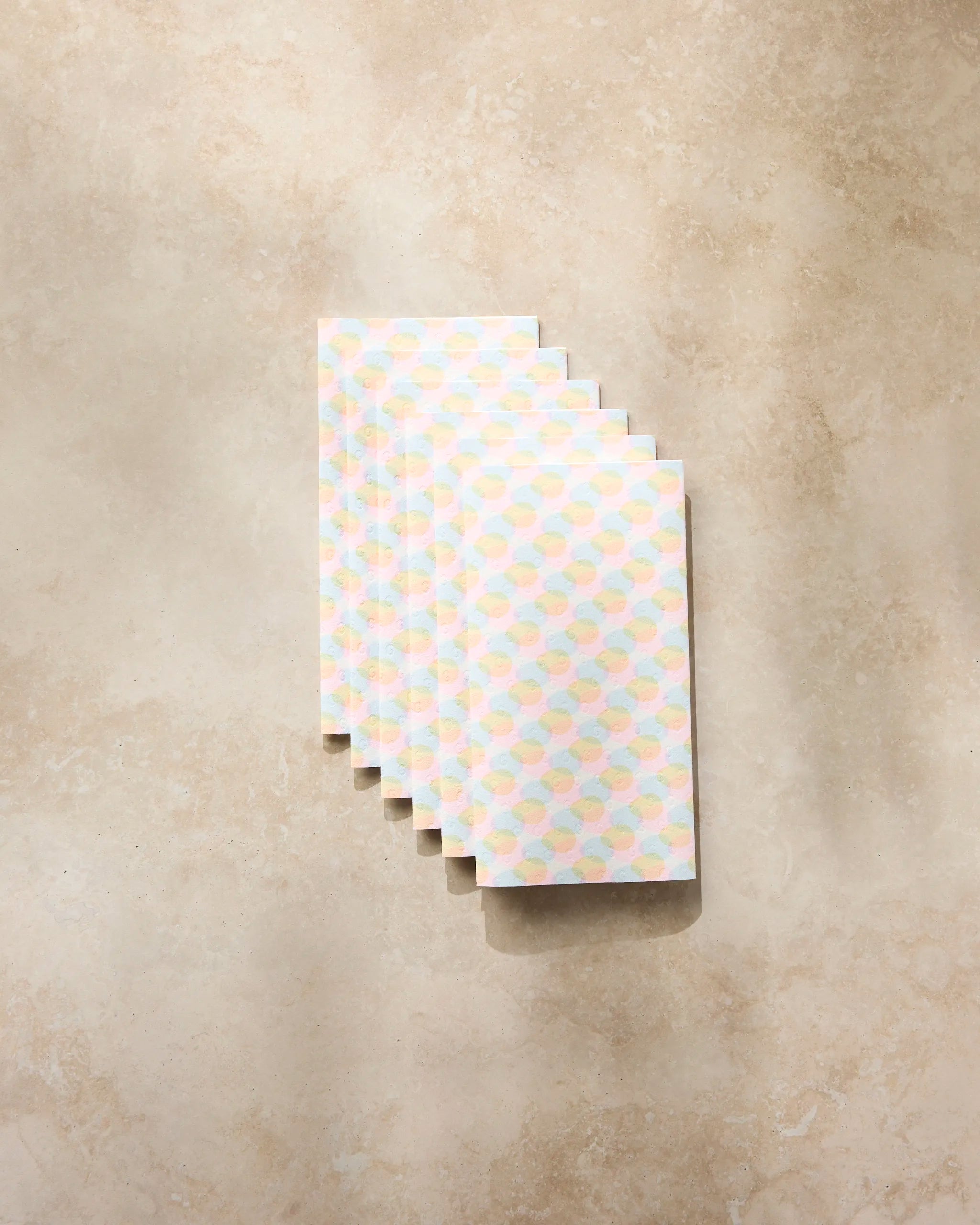 Six Geometry Pastel Pop small notebooks, featuring geometric pastel patterns, are stacked on a beige marble surface, their soft hues highlighted by sunlight and subtle shadows.