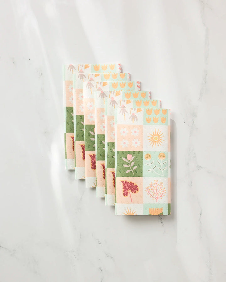 Six "Patchwork Garden" notebooks by Rachel Parker, with pastel covers adorned with floral and sun designs, are stacked neatly on white marble—a stylish and eco-friendly choice for fans of recycled products or reusable paper towels.