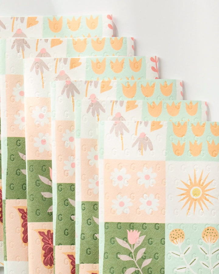 Rachel Parker's Patchwork Garden features eco-friendly, pastel kitchen towels in a fan arrangement, each with patchwork squares displaying floral and sun designs in green, yellow, pink, and beige.