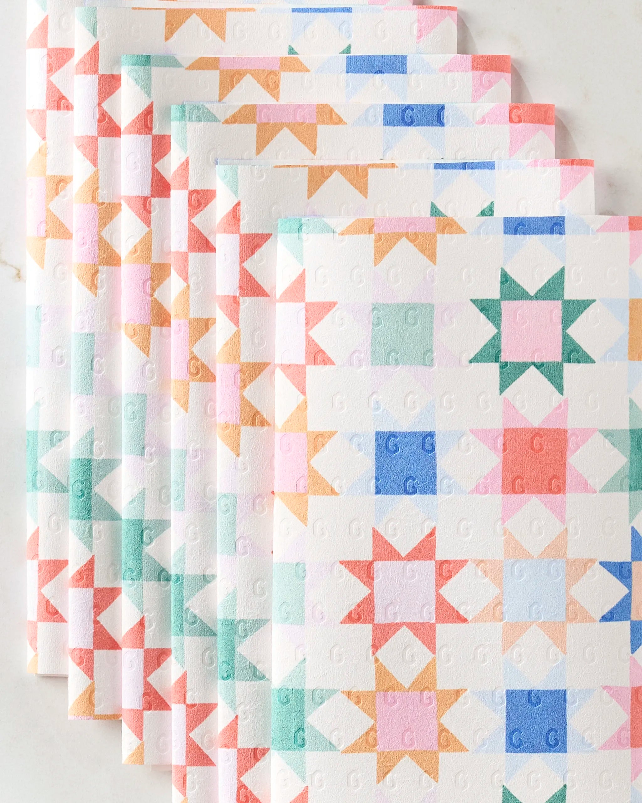 The Patchwork Picnic kitchen towels by Brittany Jepsen, featuring eco-friendly pastel fabric with a geometric star and square quilt pattern, are neatly stacked in an overlapping display on a light surface.