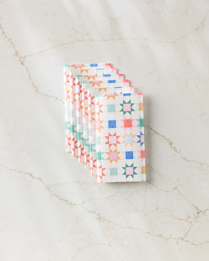 Six Patchwork Picnic kitchen towels by Brittany Jepsen, featuring a colorful geometric star pattern, are neatly folded and stacked on a light marble surface.