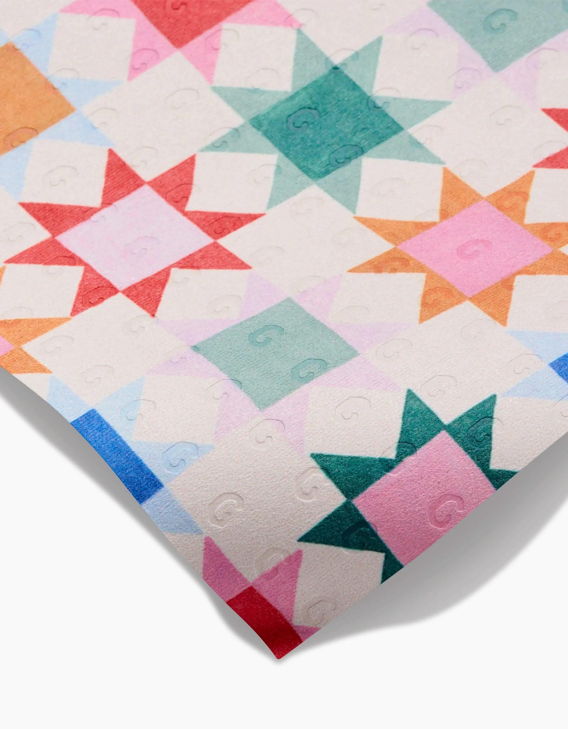 A close-up of the Patchwork Picnic quilt by Brittany Jepsen, showcasing a geometric star pattern in vibrant red, green, pink, orange, and blue on cream. Made from eco-friendly fabric with a subtle embossed G design.