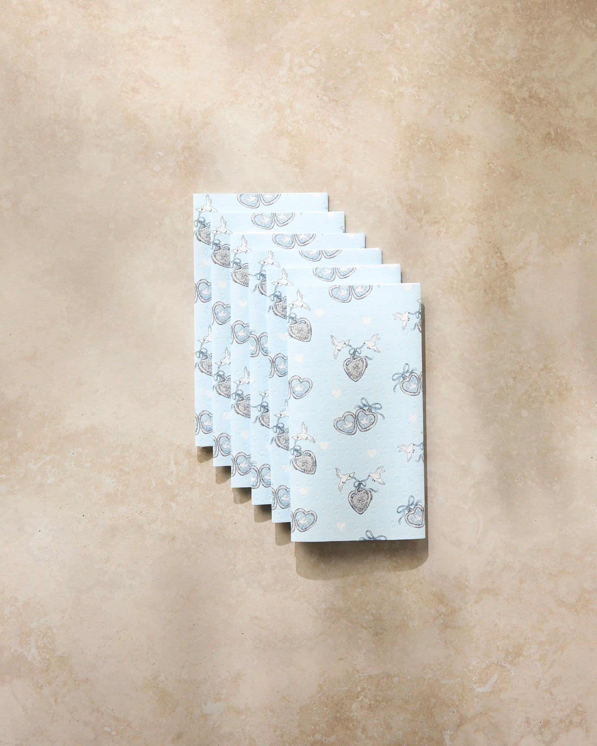 Six rectangular Peace Doves bars by Geometry, wrapped in light blue eco-friendly paper towels with heart, bow, and key designs, are neatly arranged on a beige stone surface.