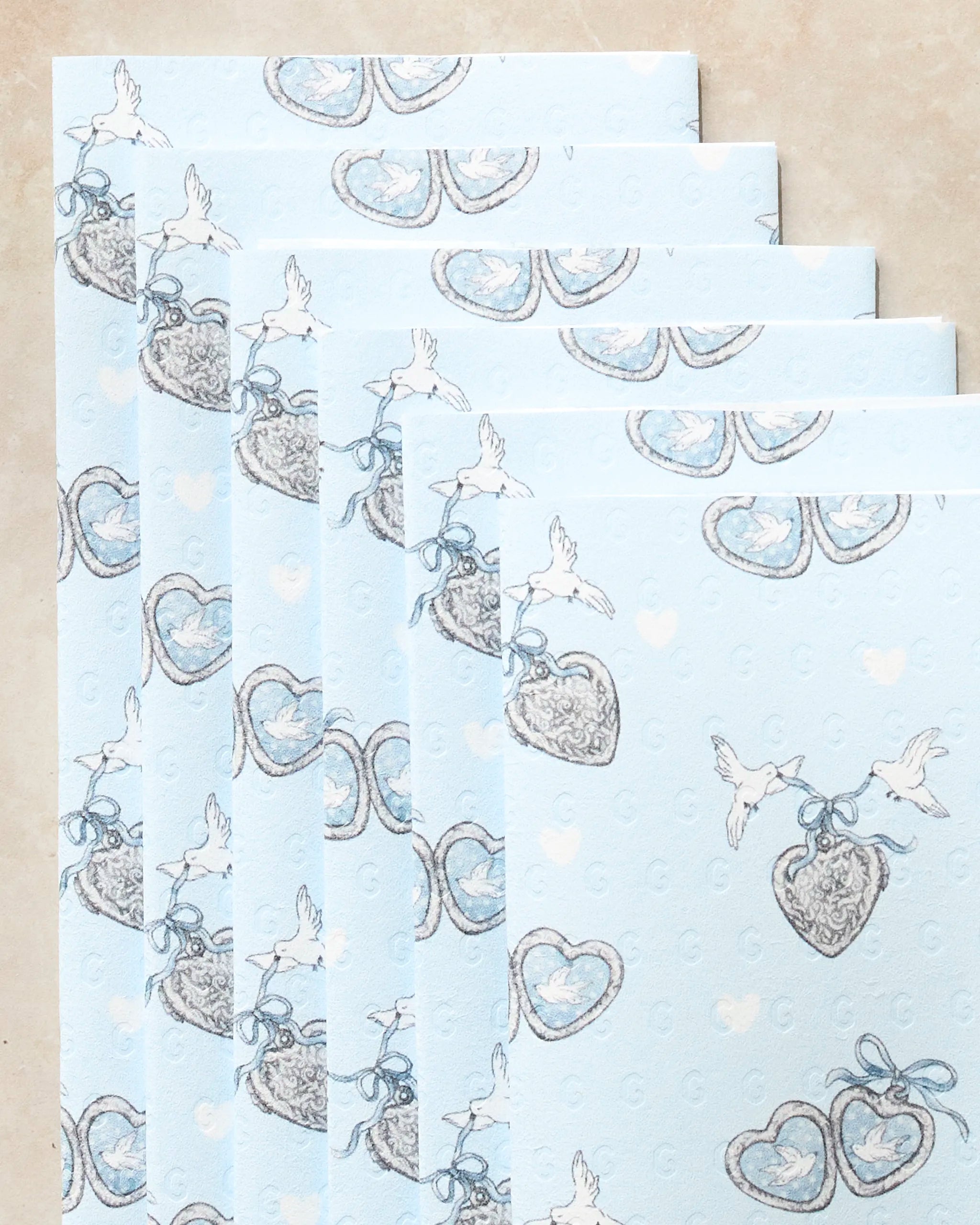 Five light blue Geometry Peace Doves eco-friendly paper towels, each patterned with doves holding heart-shaped lockets, are arranged in an overlapping fan shape on a beige surface.