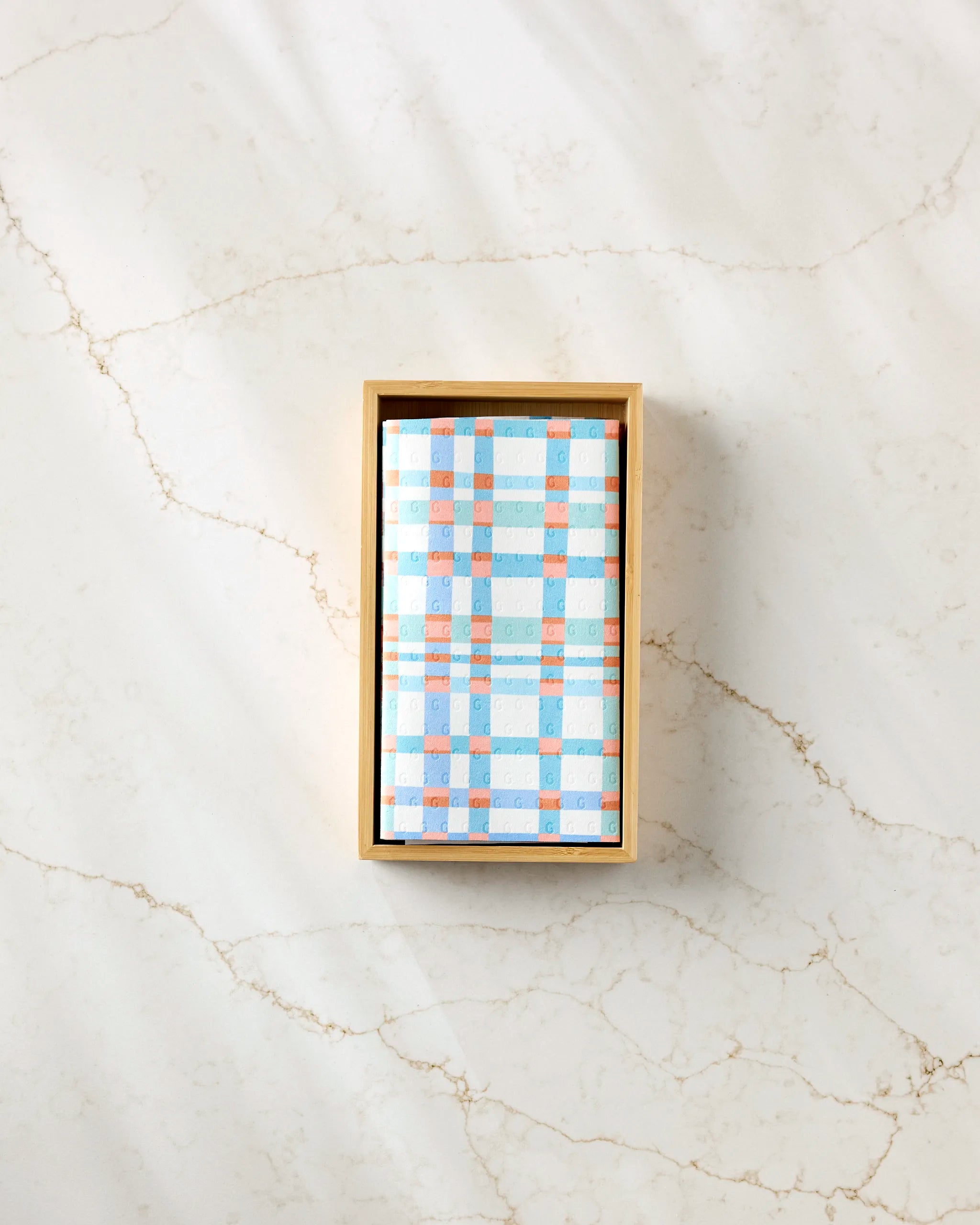 A rectangular box wrapped in Rachel Parker's Picnic Perfect blue, white, and orange plaid reusable paper towels sits on a light marble surface with subtle veins.