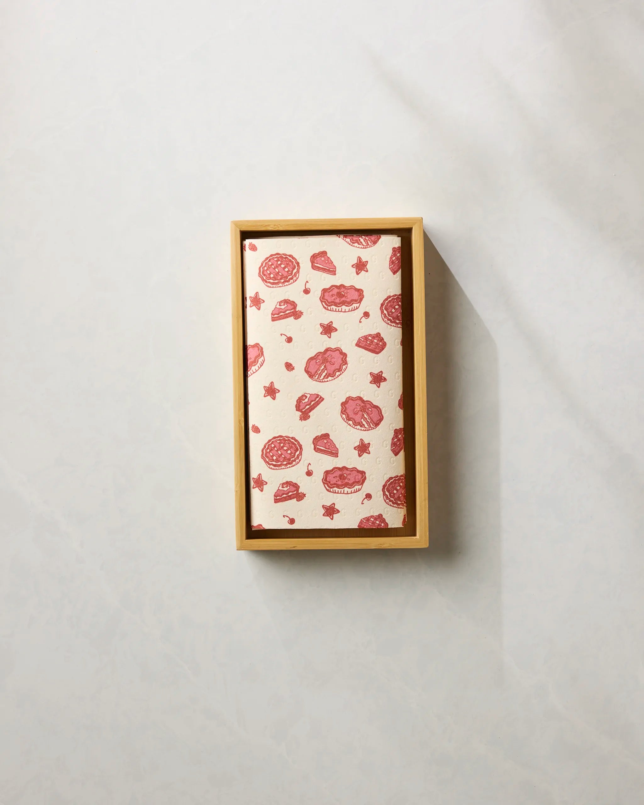 The Pie-triotic box by Geometry, featuring a pink and red pattern of donuts, cookies, and stars on its lid, is perfect for storing reusable paper towels and sits on a light surface with a shadow to the right.