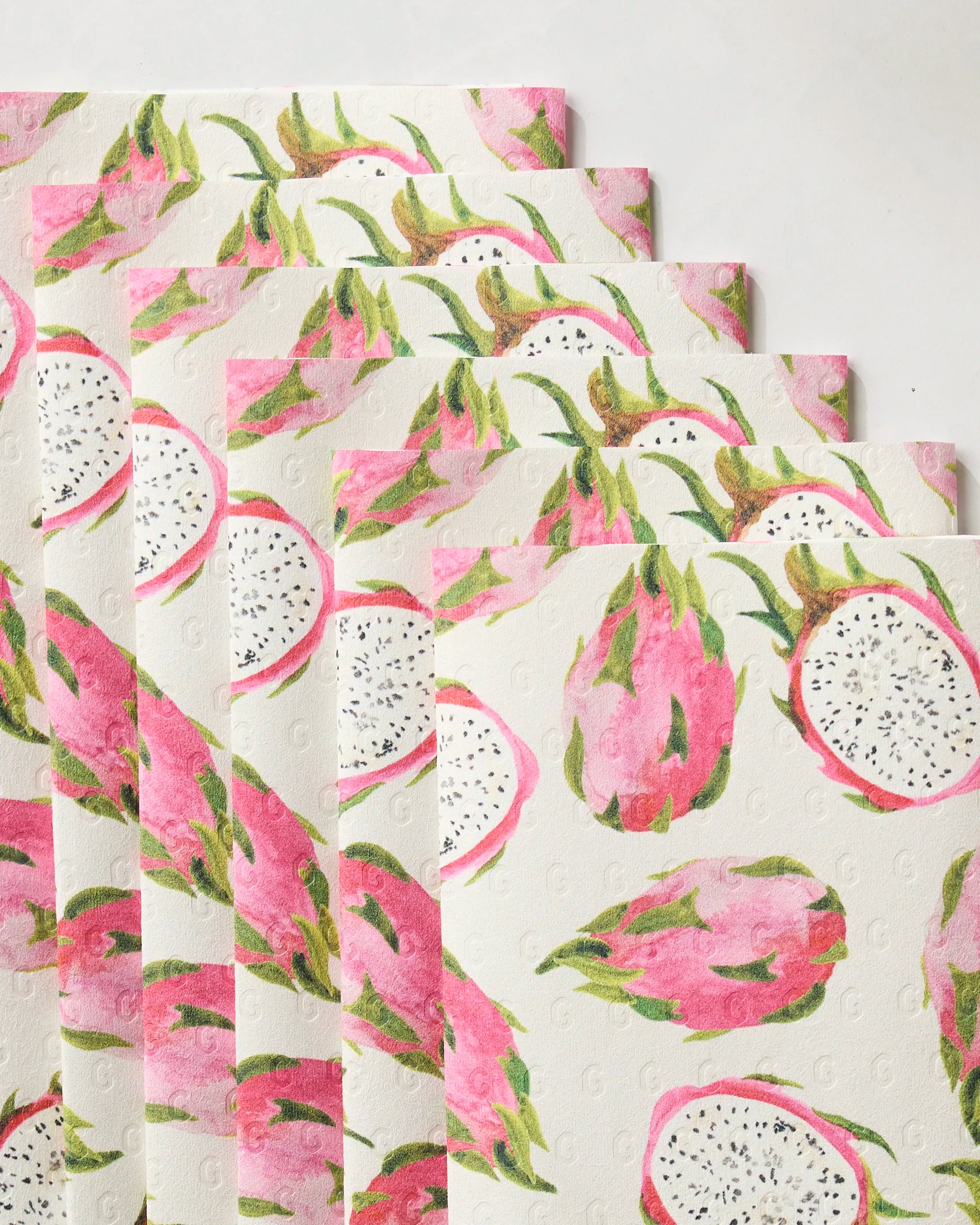 Several square, eco-friendly Geometry Pink Dragon kitchen towels, featuring pink, green, and white dragon fruit illustrations, are arranged in a fanned-out stack on a white surface.