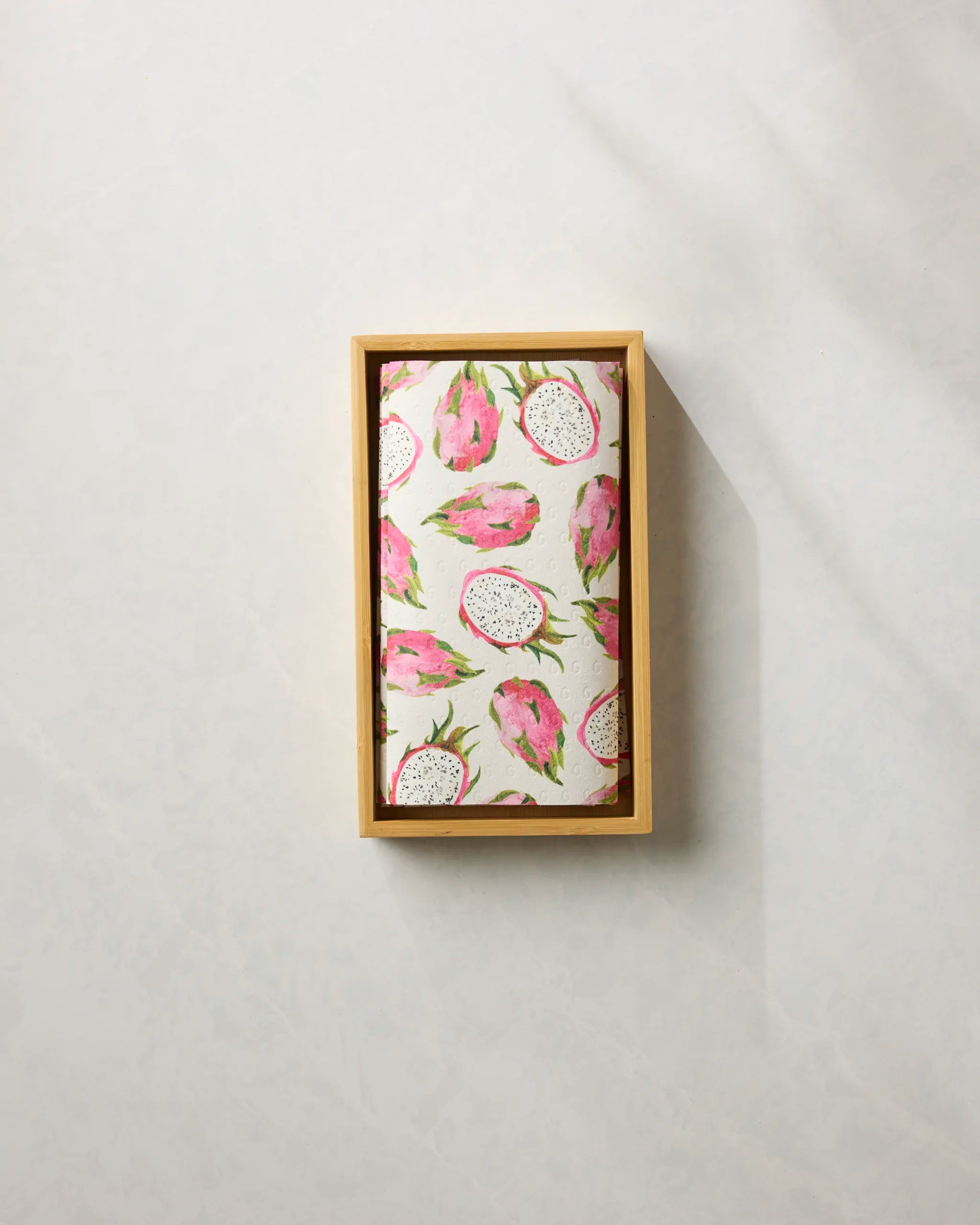 A Pink Dragon kitchen towel by Geometry, featuring a dragon fruit pattern and eco-friendly design, is neatly folded in a wooden box against a neutral background.