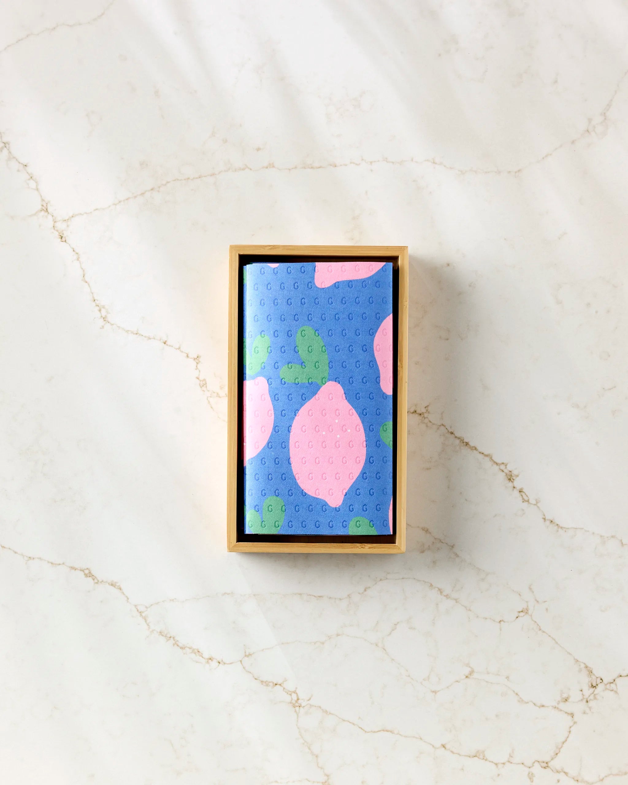 A rectangular box contains Pink Lemons reusable paper towels by Jennifer Bouron—a folded blue cloth with a pink lemon and green leaf pattern—displayed on a light marble surface.