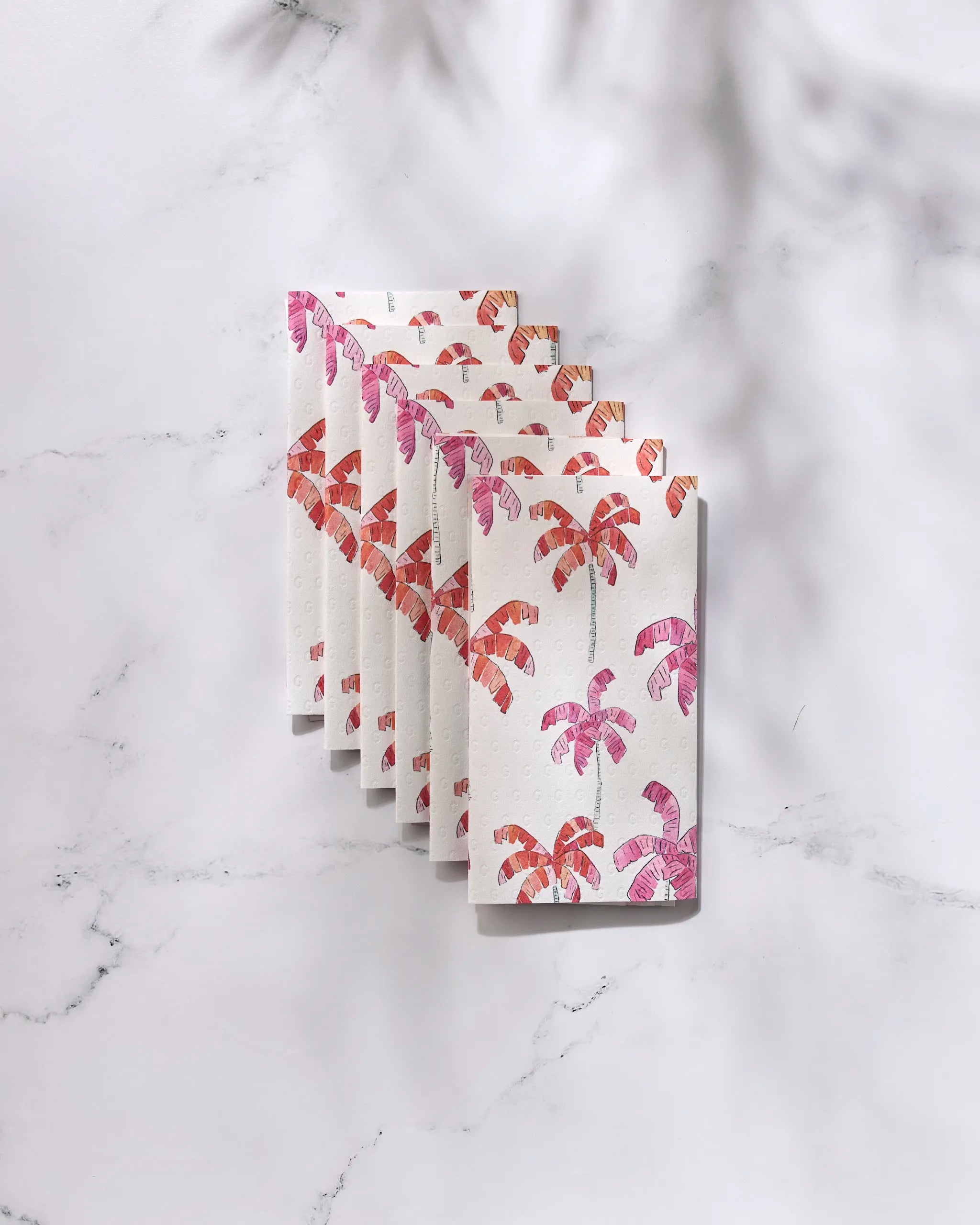 Six GEOMETRY "Pink Palms" greeting cards, featuring pink and orange palm tree patterns, are fanned out on white marble with palm leaf shadows above—each crafted with sustainable, eco-friendly paper towels.