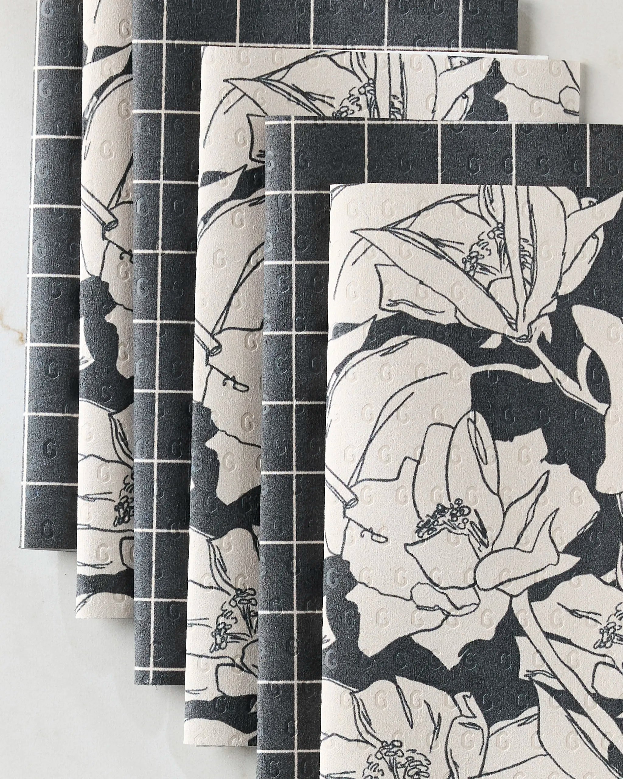 Four overlapping "Plant One on Me" notebooks by Aubrey Fairchild, featuring dark grid covers with bold white florals, arranged on a light surface—an ideal match for your eco-friendly kitchen or reusable paper towels.