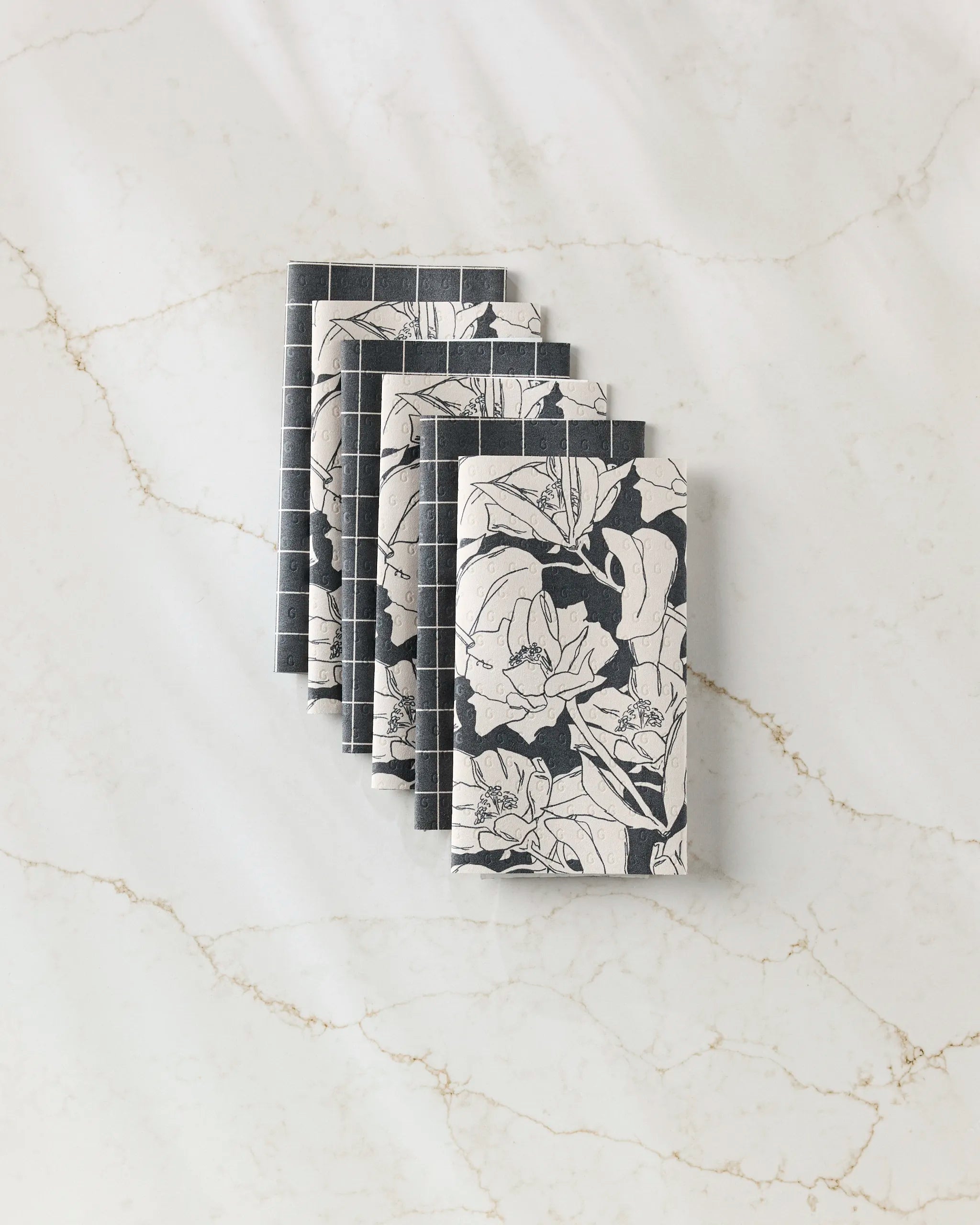 Six “Plant One on Me” napkins by Aubrey Fairchild, featuring a floral black-and-white design, are eco-friendly kitchen towels arranged in a staggered row on a light marble-textured surface.
