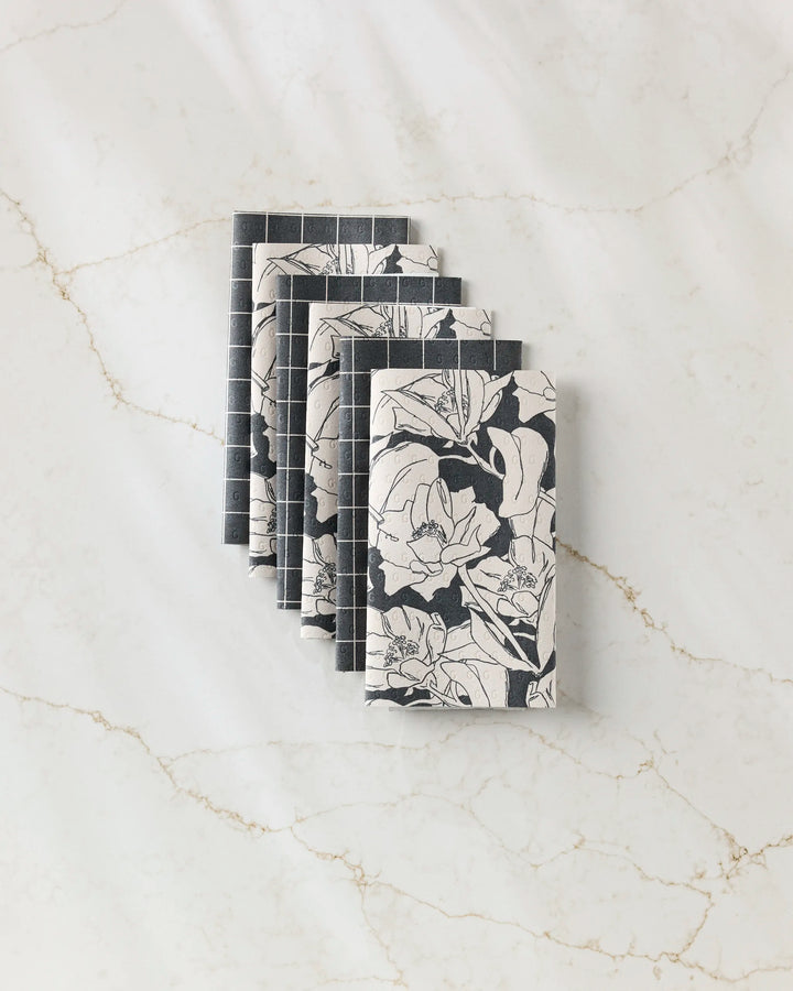 Six “Plant One on Me” napkins by Aubrey Fairchild, featuring a floral black-and-white design, are eco-friendly kitchen towels arranged in a staggered row on a light marble-textured surface.