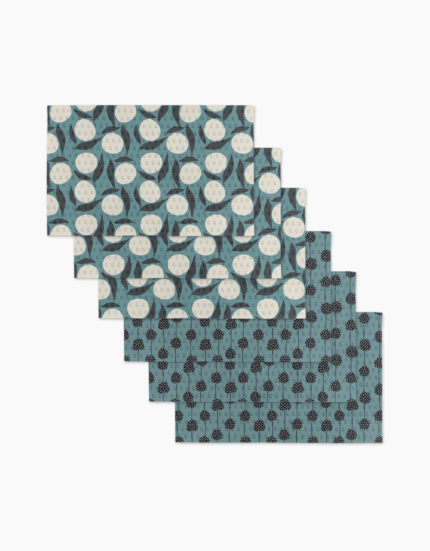 Six Polka Dot Blooms placemats by Lizzie Clark are arranged in a staggered stack: three have white circles with curved blue shapes, and three feature black floral motifs on a blue background. Made from reusable paper towels.