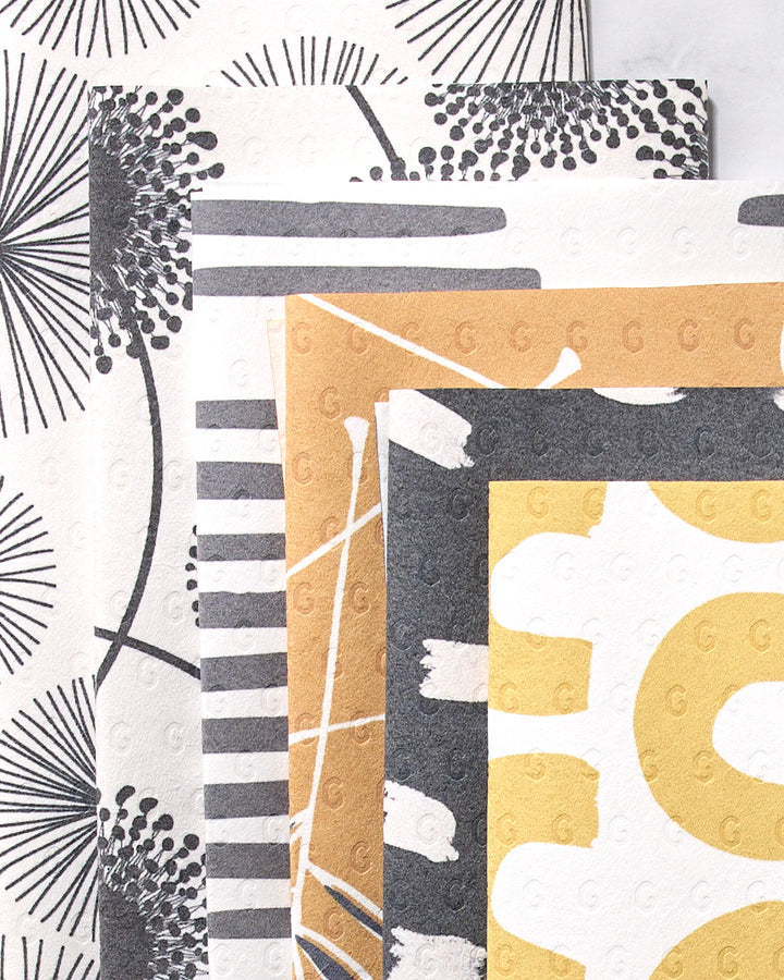 Four GEOMETRY Pop Of Color reusable towels, featuring bold modern patterns in black, white, gold, and gray with abstract florals, stripes, and brush strokes designs, are shown overlapping. Each towel has a subtle embossed texture.