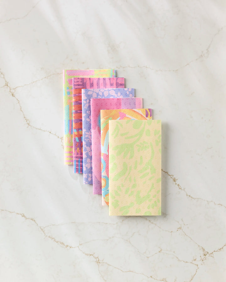 A row of Sareka Unique’s Positive Vibes notebooks, each with a unique pastel abstract design, is neatly arranged on a white marble surface, their patterns reminiscent of eco-friendly kitchen towels.