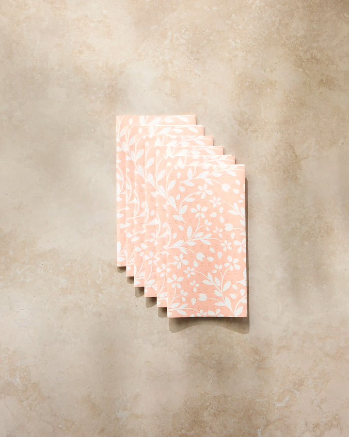 A stack of six Quiet Radiance cards by Geometry, featuring a white floral pattern on pink, arranged on a light beige textured surface—evoking the elegance and sustainability of reusable paper towels.
