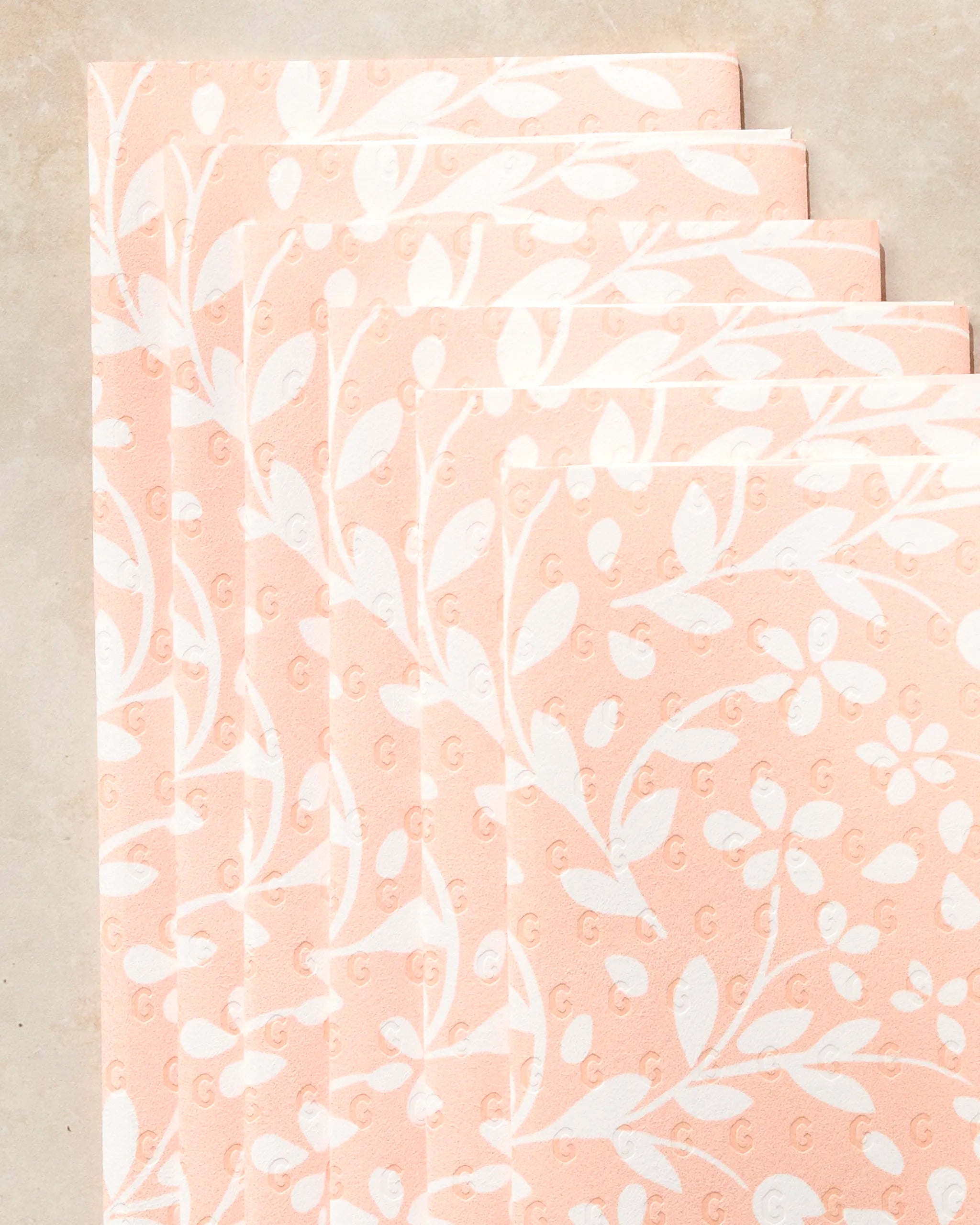 Five peach-colored Quiet Radiance paper towels by Geometry, featuring a white floral and leaf pattern, arranged in an overlapping diagonal stack on a light beige surface.