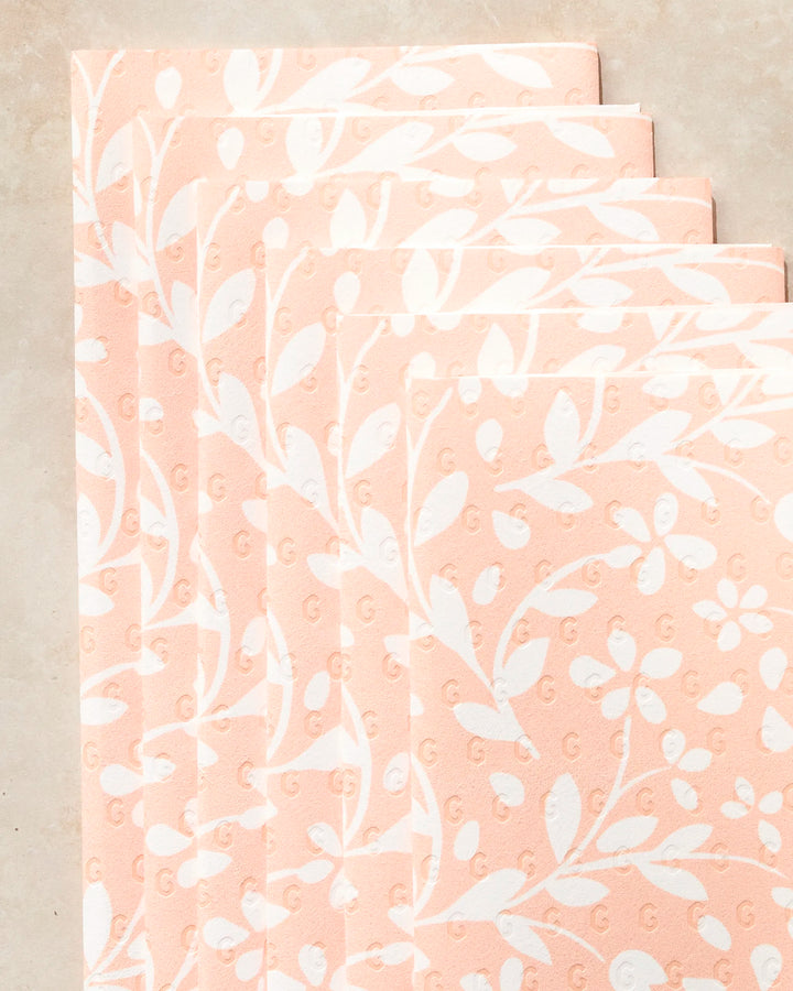 Five peach-colored Quiet Radiance paper towels by Geometry, featuring a white floral and leaf pattern, arranged in an overlapping diagonal stack on a light beige surface.
