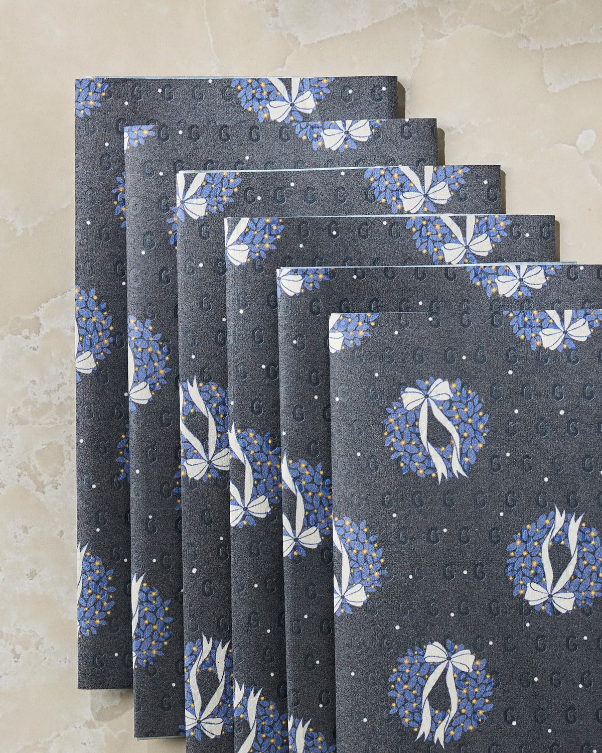 Six folded sheets of Geometry’s Quiet Skies reusable paper towels, featuring dark gray hues with blue and white wreath patterns and subtle “G” motifs, are arranged in a fan shape on a light marble surface. These eco-friendly alternatives to traditional kitchen towels offer an elegant touch while promoting sustainability for your home.