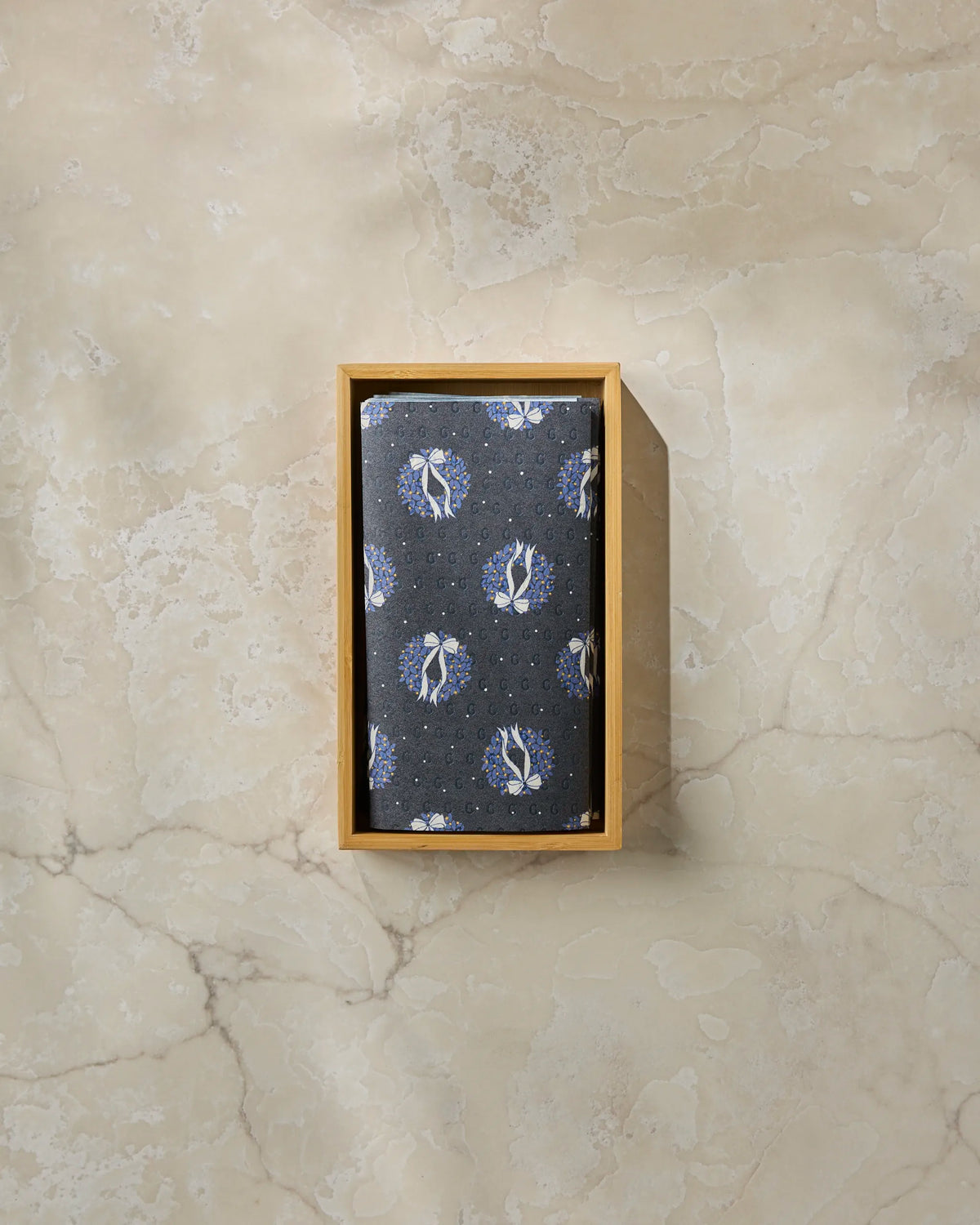 A neatly folded “Quiet Skies” dark floral-patterned eco-friendly kitchen towel by Geometry rests inside a rectangular wooden box on a light marble surface.