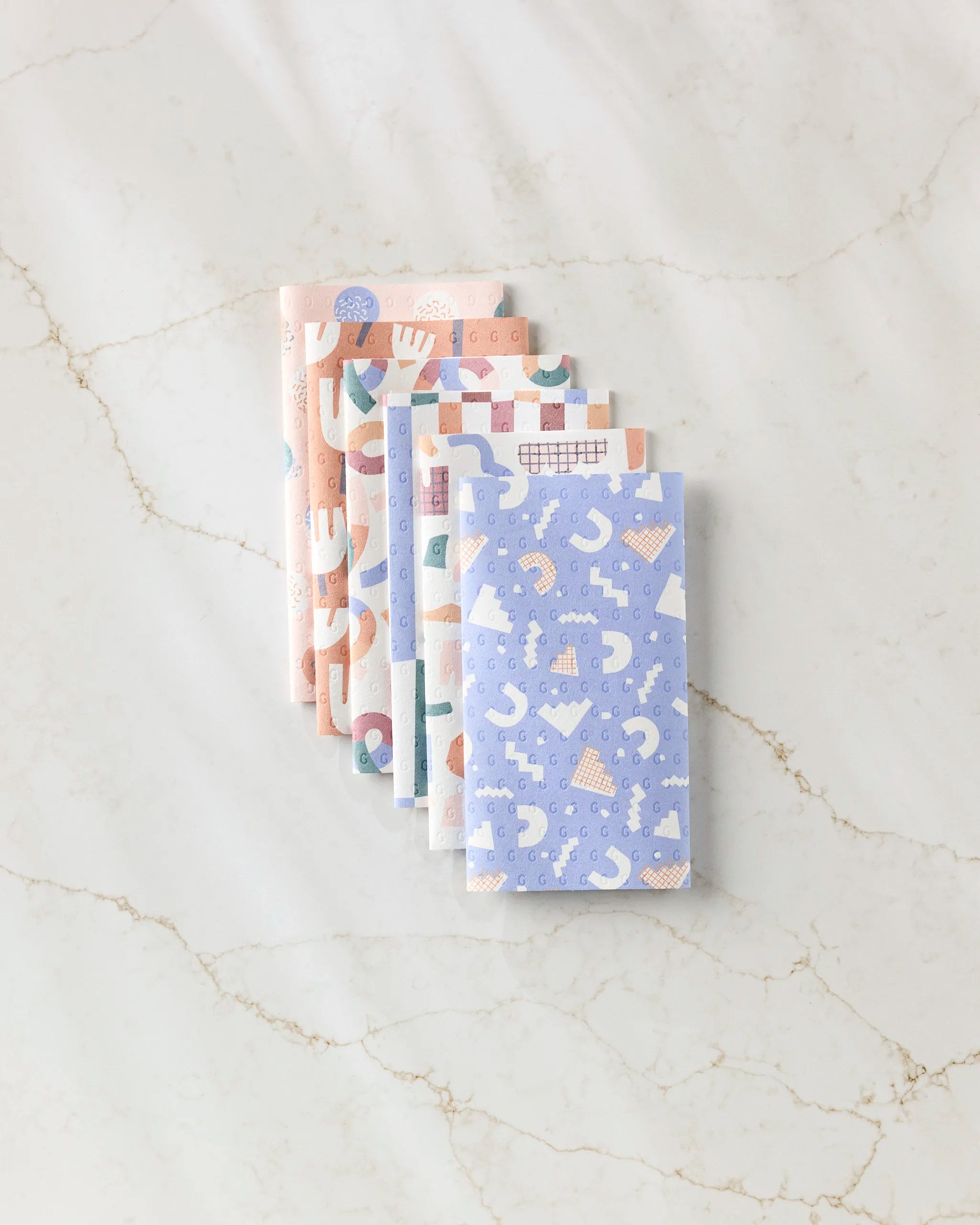 Six Rainbow Cutouts reusable paper towels by Claire Iglesias, featuring pastel colors and playful abstract patterns, are stacked neatly on a white marble-like surface with subtle veining.