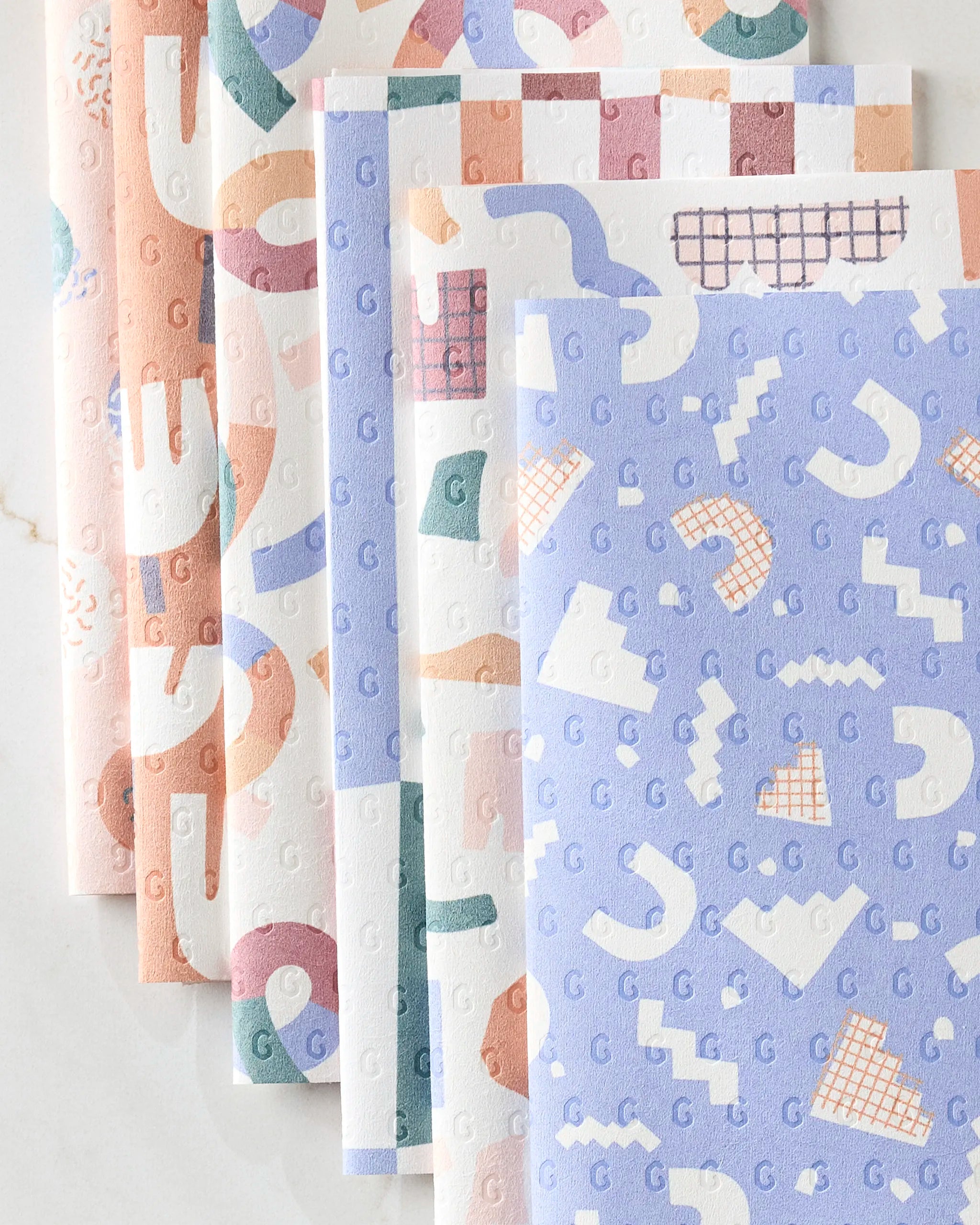 Claire Iglesias’ Rainbow Cutouts features several overlapping sheets of patterned, reusable paper towels in pastel colors with abstract geometric shapes, squiggles, and repeating letter G designs displayed on a light surface.