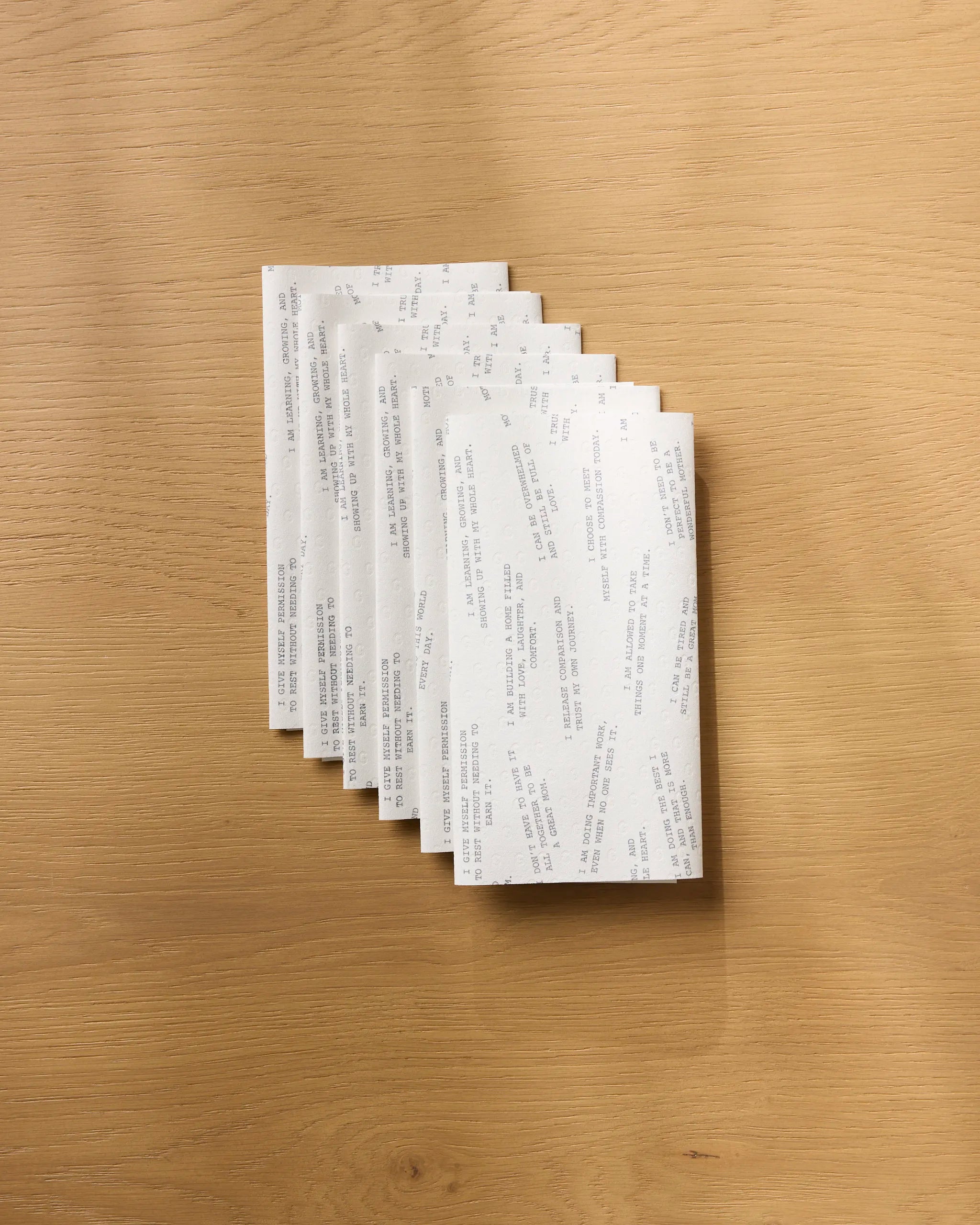 Seven Reminders for Mom alcohol prep pads by Geometry are fanned out on a light wooden surface. Their white, grey-printed wrappers sit next to a stack of eco-friendly paper towels.
